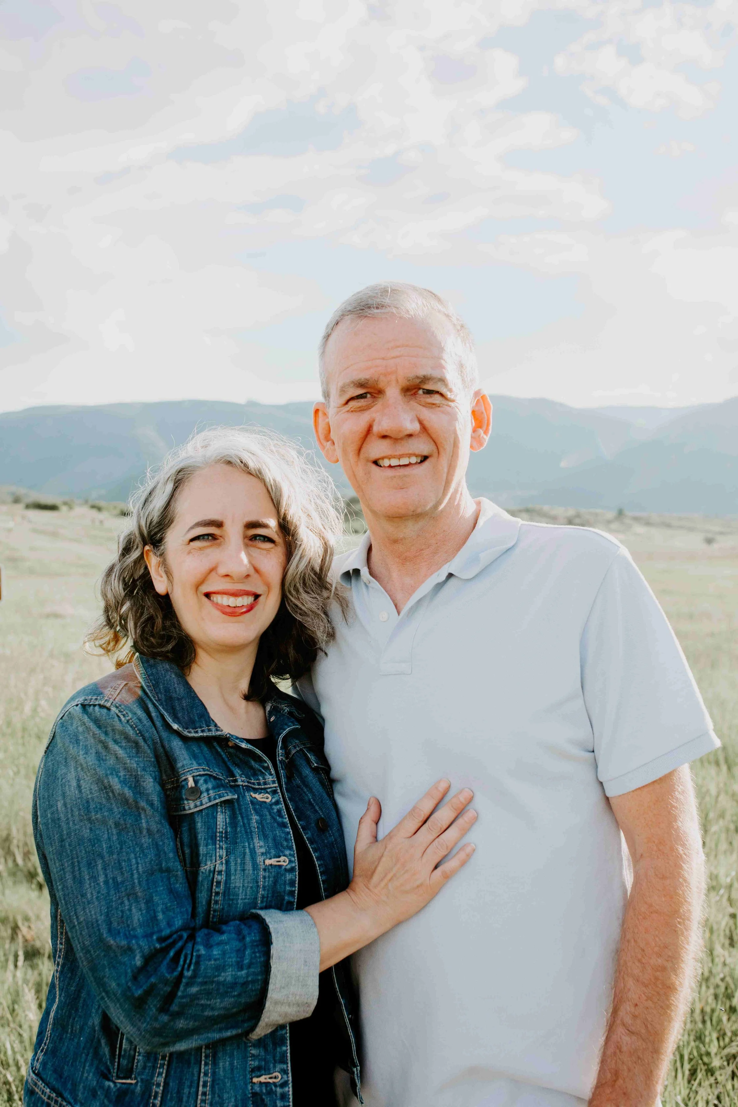 A woman in a denim jacket and a man in a light polo shirt standing together in a grassy field with mountains in the background.