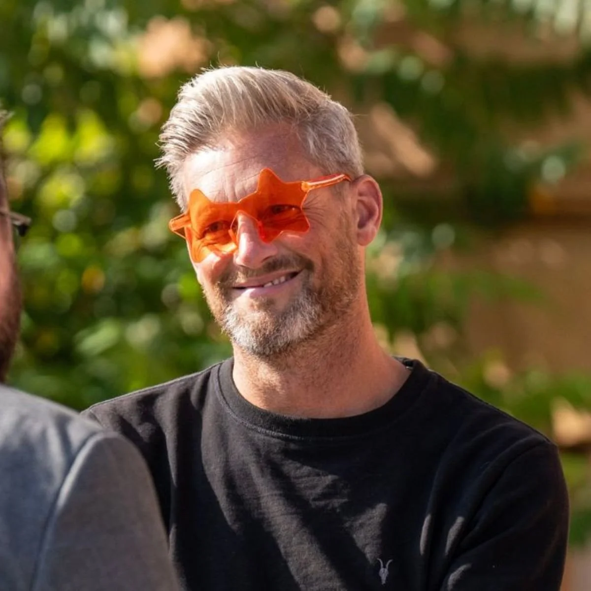 A smiling man with gray hair and a beard wearing orange star-shaped sunglasses outdoors surrounded by green foliage.