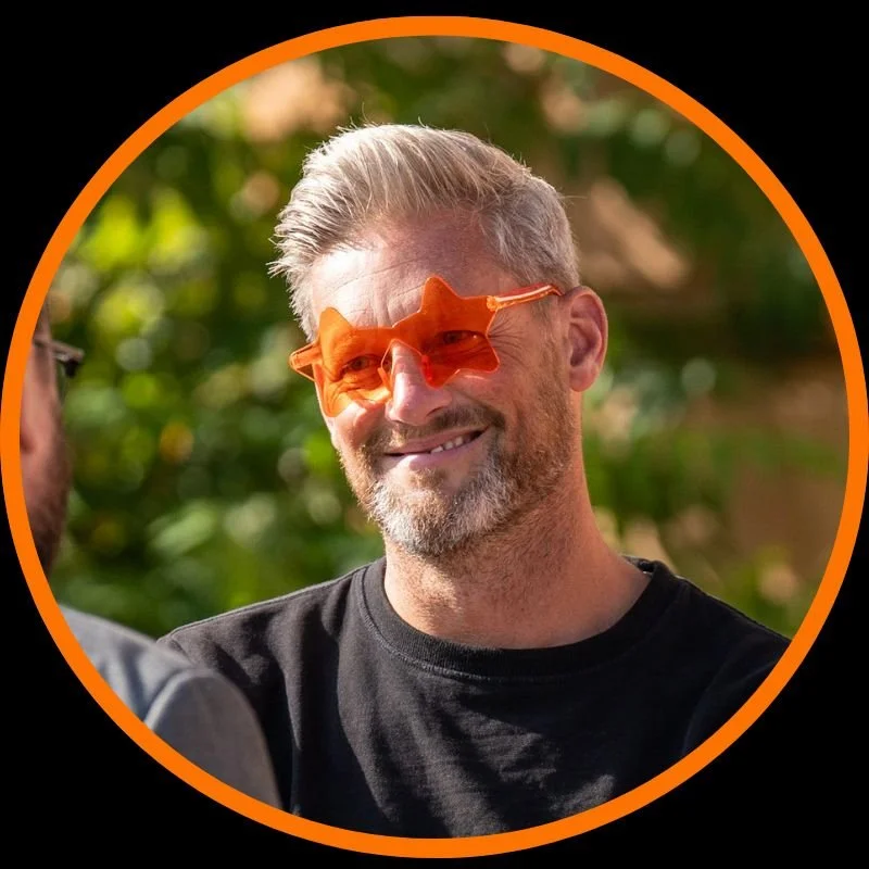 Smiling man with gray hair and beard wearing orange novelty sunglasses outdoors.