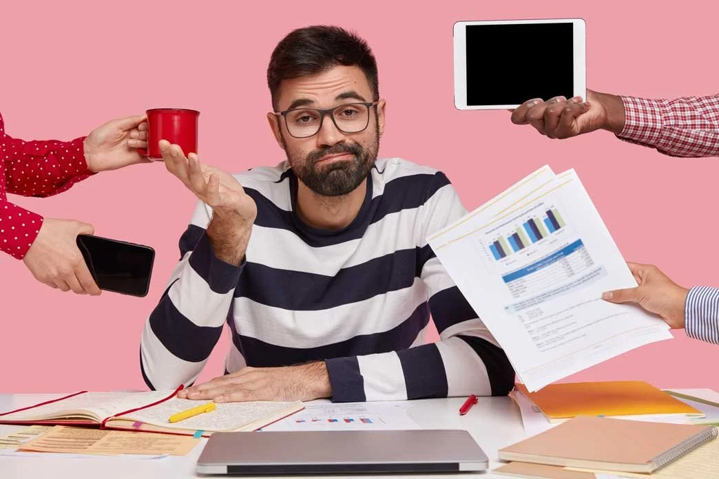 Man with glasses in a striped shirt surrounded by hands offering various objects, including a tablet, a smartphone, a coffee mug, and documents, in front of a pink background.