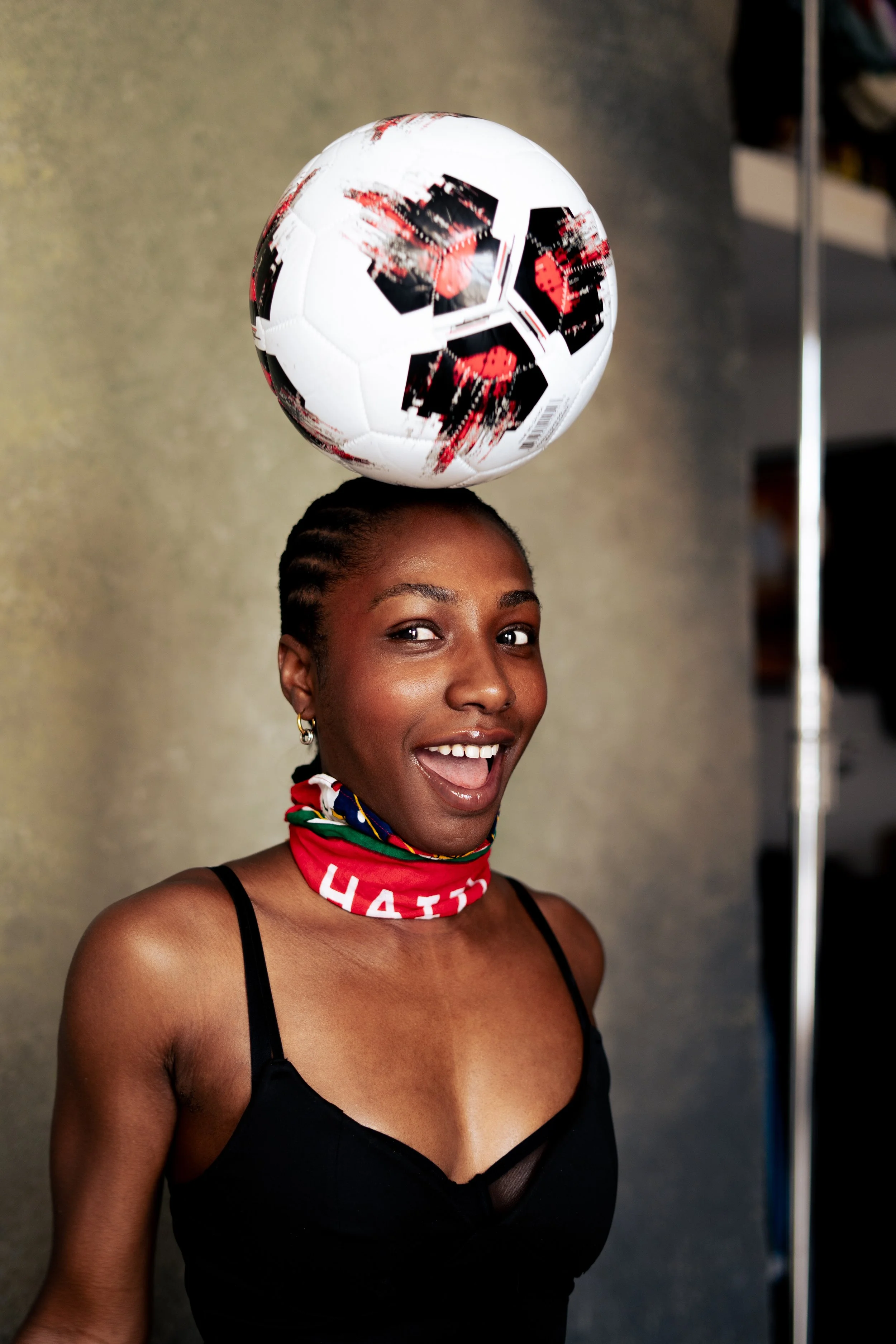 A young woman with dark hair in braids, smiling and balancing a soccer ball on her head. She is wearing a black spaghetti strap top and a red scarf with white text around her neck. The background is a plain wall.