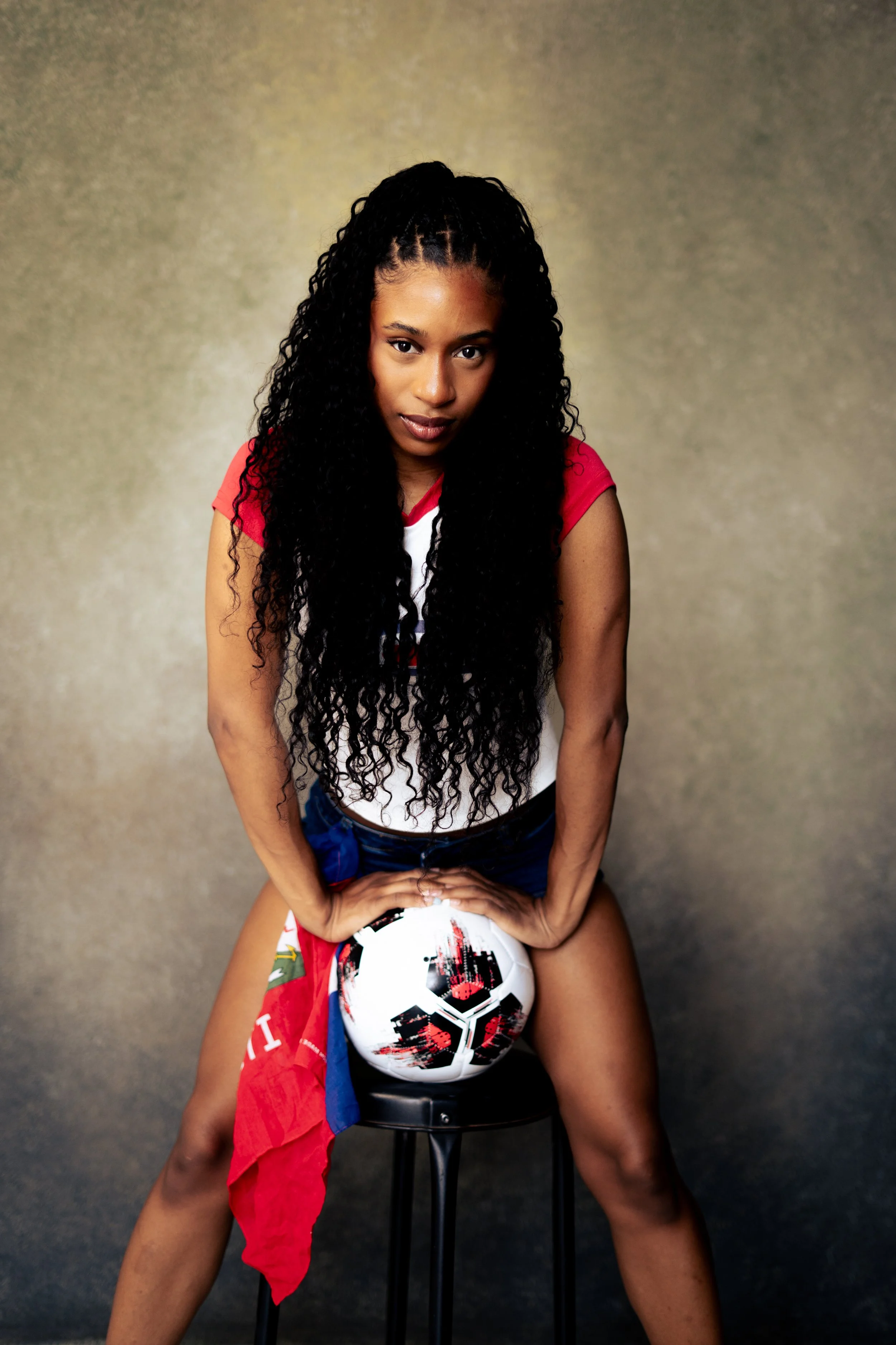 A young woman with long, curly black hair and dark eyes, wearing a red t-shirt, sits on a stool. She is leaning forward with her hands on her thighs, over a soccer ball, and looking directly at the camera. The background is a plain, textured beige wa