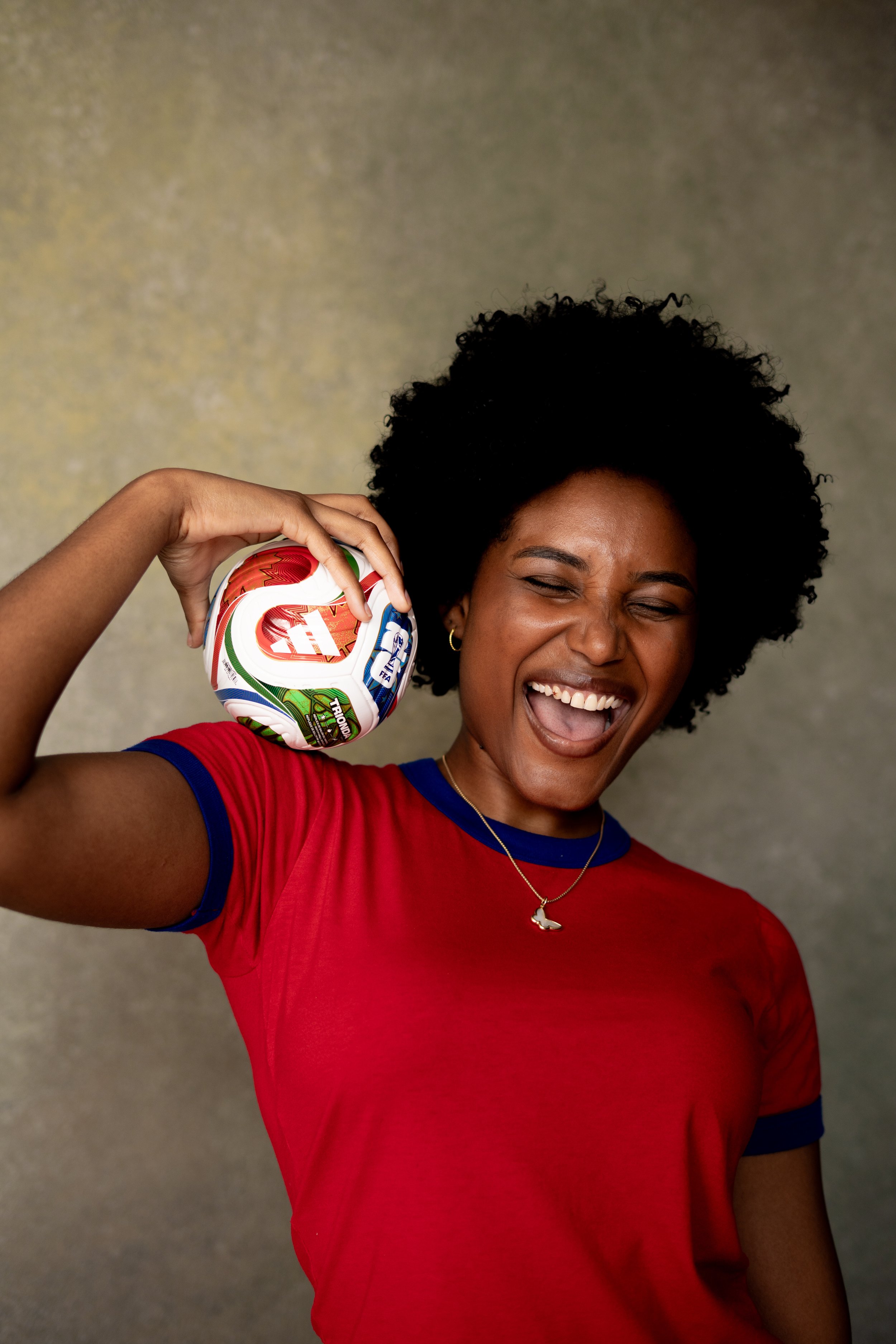 A woman with curly black hair wearing a red sports jersey holding a soccer ball near her ear, smiling with her eyes closed.