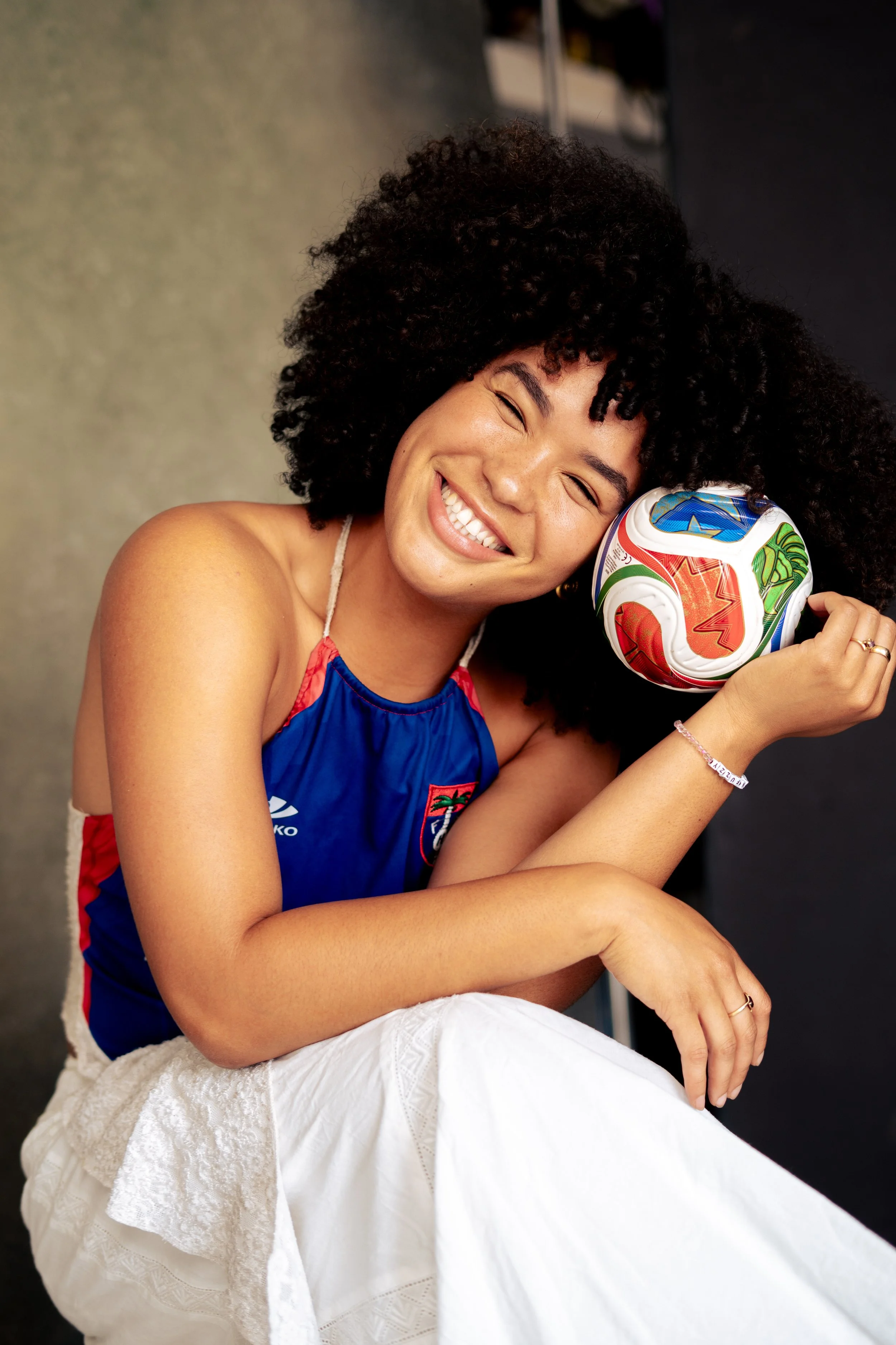 Smiling woman with curly hair, wearing a blue sports jersey, sitting on a white textured blanket, holding a colorful soccer ball close to her face.