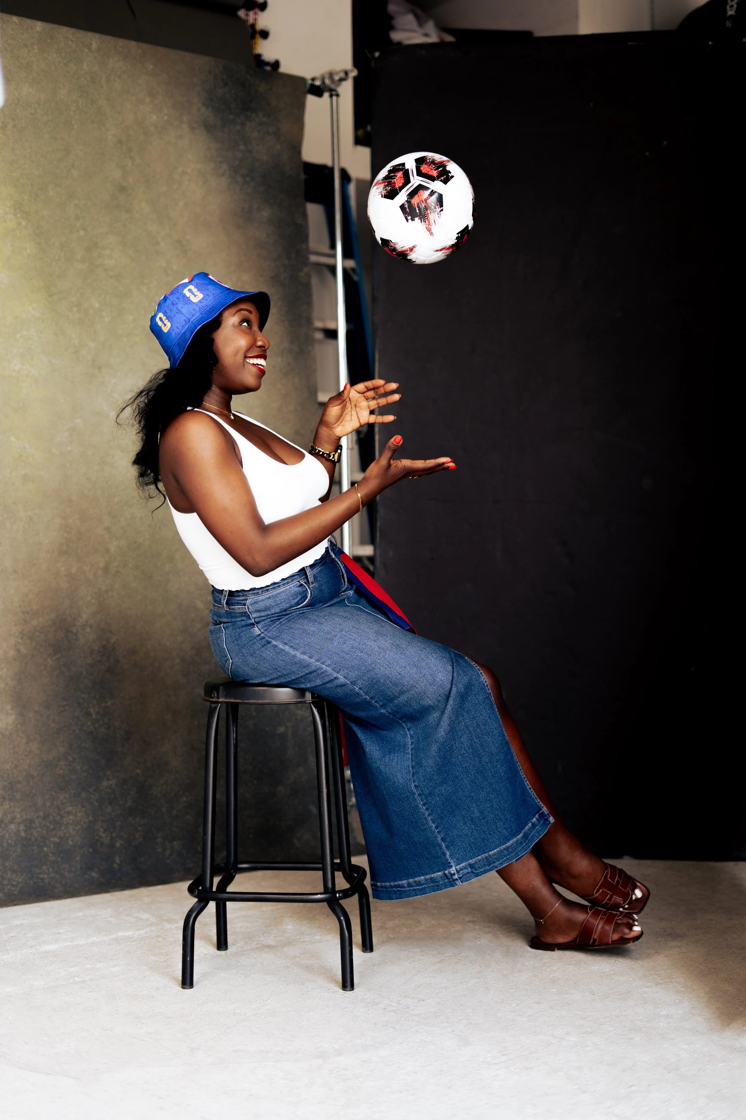 A woman sitting on a bar stool, wearing a blue bucket hat, white tank top, blue denim skirt, brown sandals, and gold jewelry, is smiling and looking at a black and white soccer ball in mid-air against a studio backdrop.