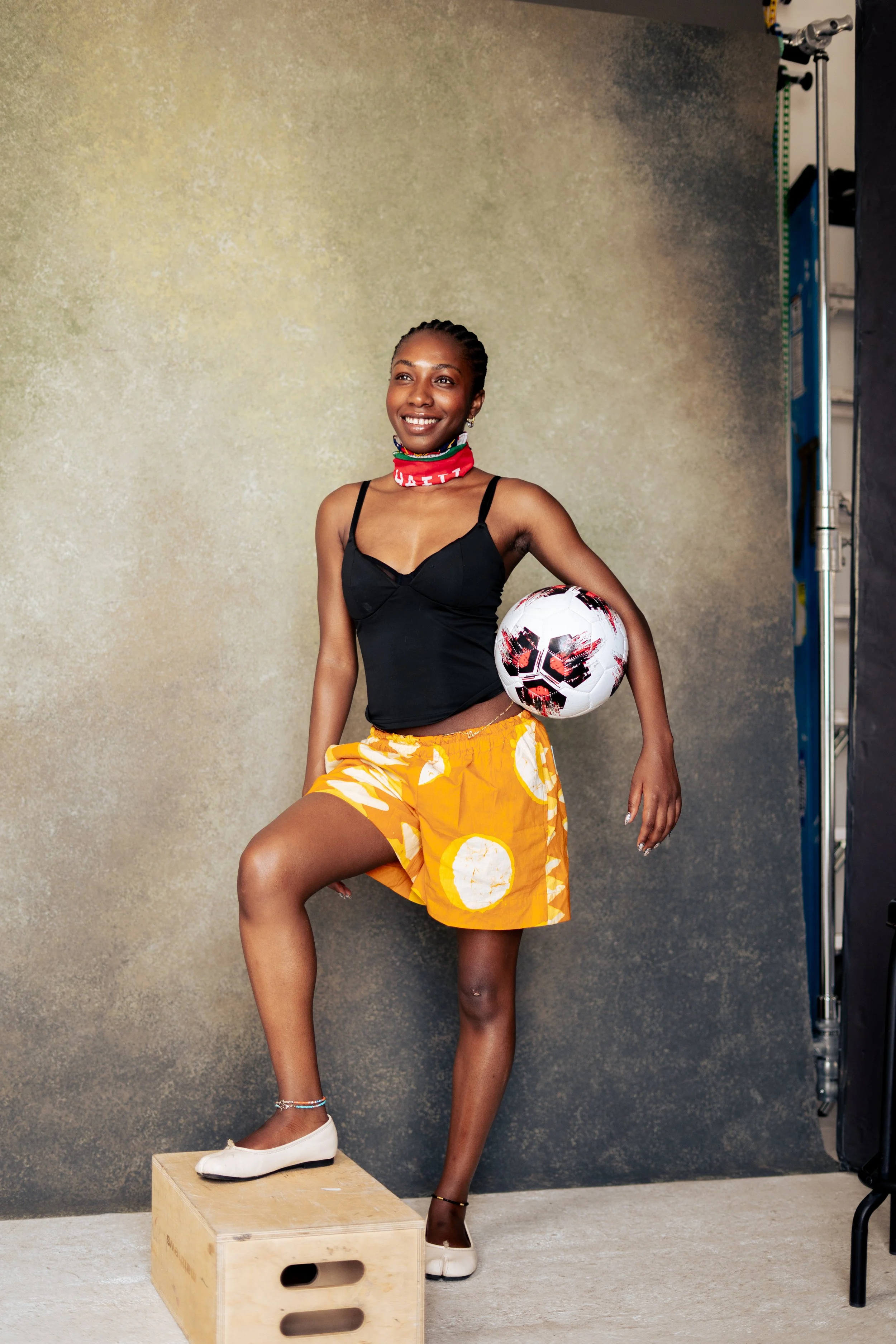 A woman with braids is standing with one foot on a wooden box, holding a soccer ball against her hip, in front of a textured gray backdrop, wearing a black top, yellow shorts with white patterns, white shoes, a red neck gaiter, and bracelets.