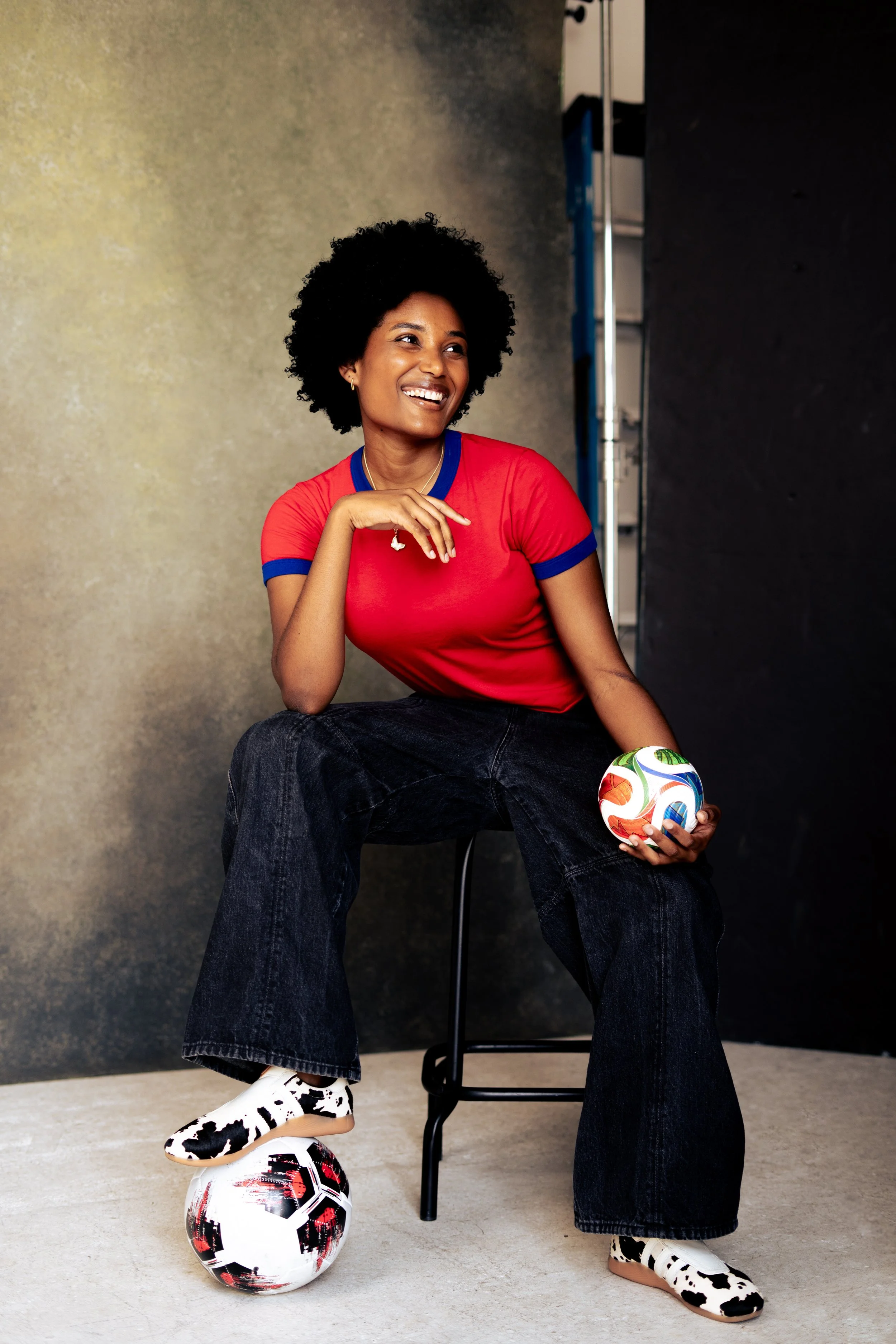 A woman with curly black hair, wearing a red t-shirt with blue collar and sleeve cuffs, dark wide-leg jeans, and cow-patterned sneakers, sitting on a chair with one foot resting on a soccer ball and holding a colorful soccer ball in her hand, smiling