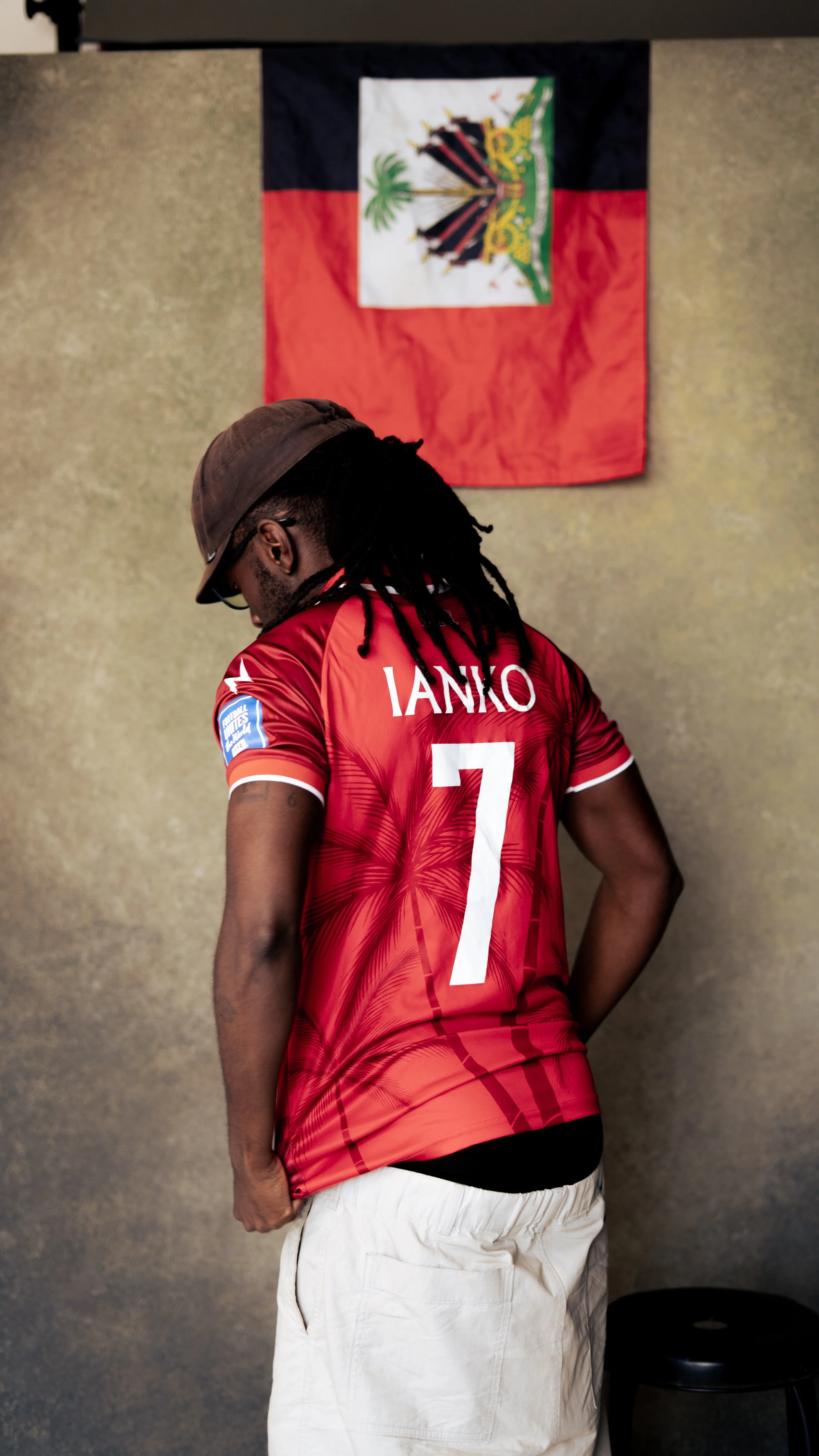 A man with dreadlocks and glasses wearing a red sports jersey with the name IANKO and the number 7 on the back, standing in front of a Tongan flag on a neutral wall background.