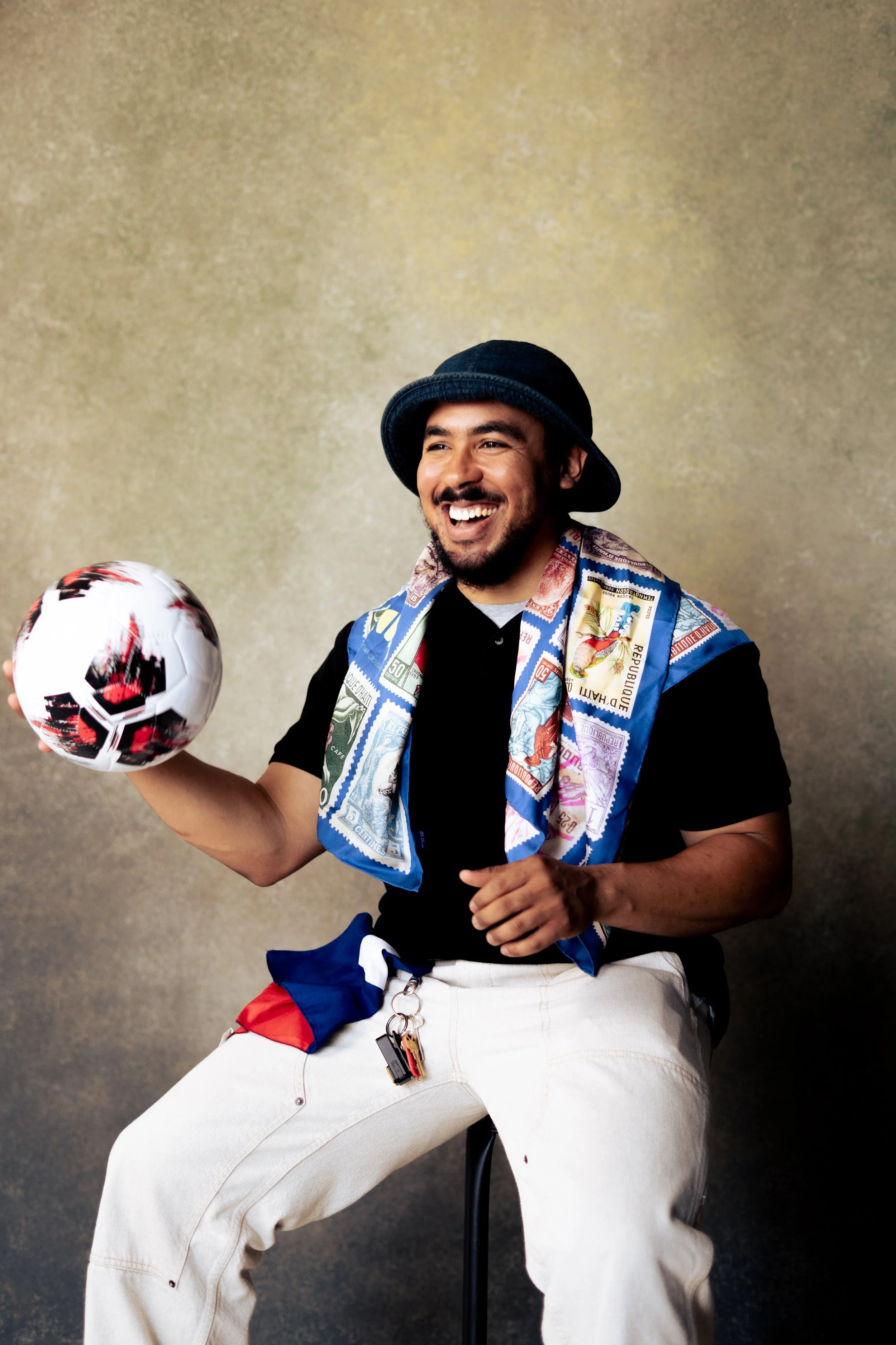 A man sitting on a chair, smiling, wearing a black hat, a colorful scarf, and white pants, holding a soccer ball, against a plain beige background.