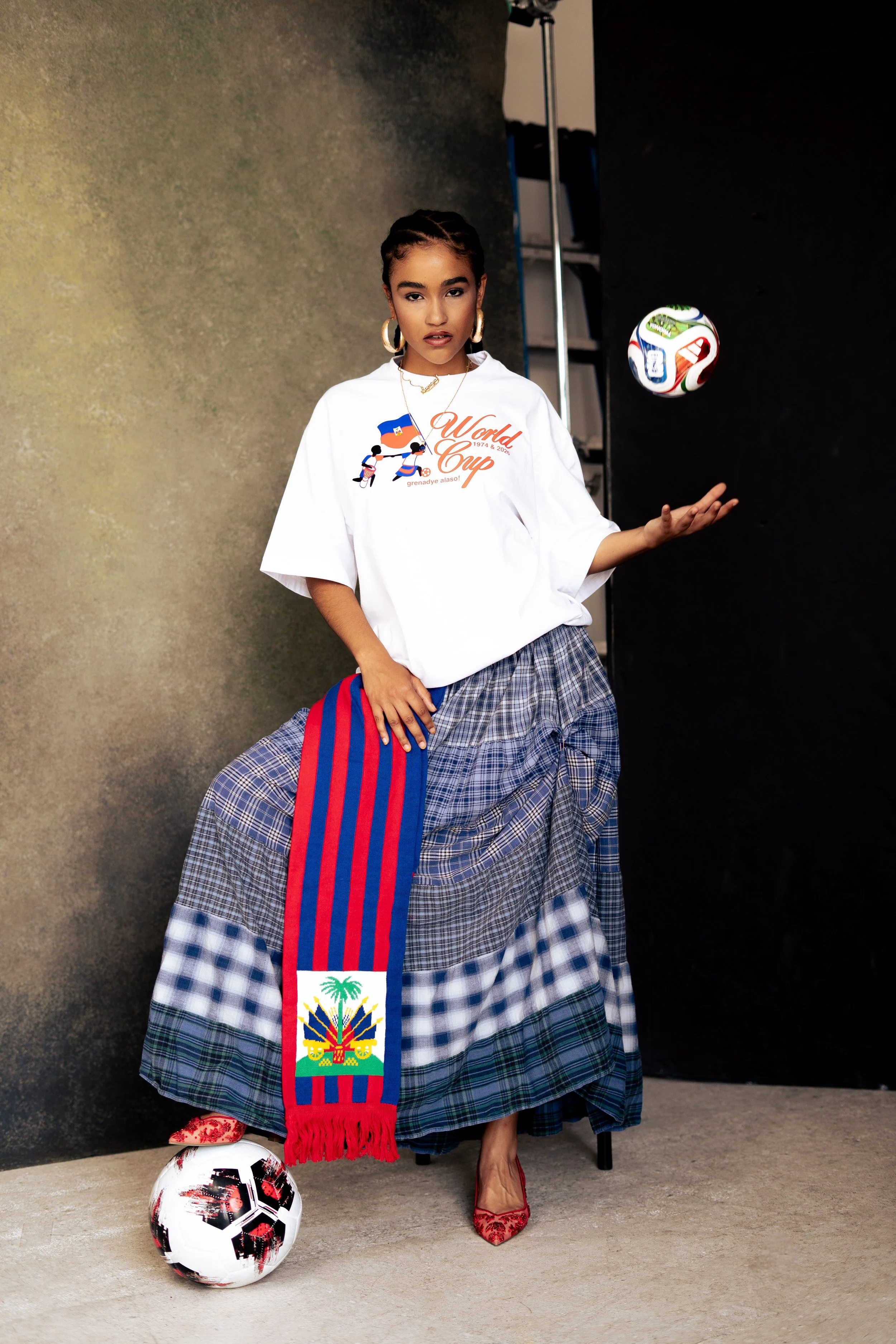A woman dressed in a white shirt with a World Cup logo, a plaid skirt, red high heels, and wearing gold jewelry, standing indoors with a soccer ball at her feet and a colorful scarf with the Haiti flag around her neck. She is positioned against a bac