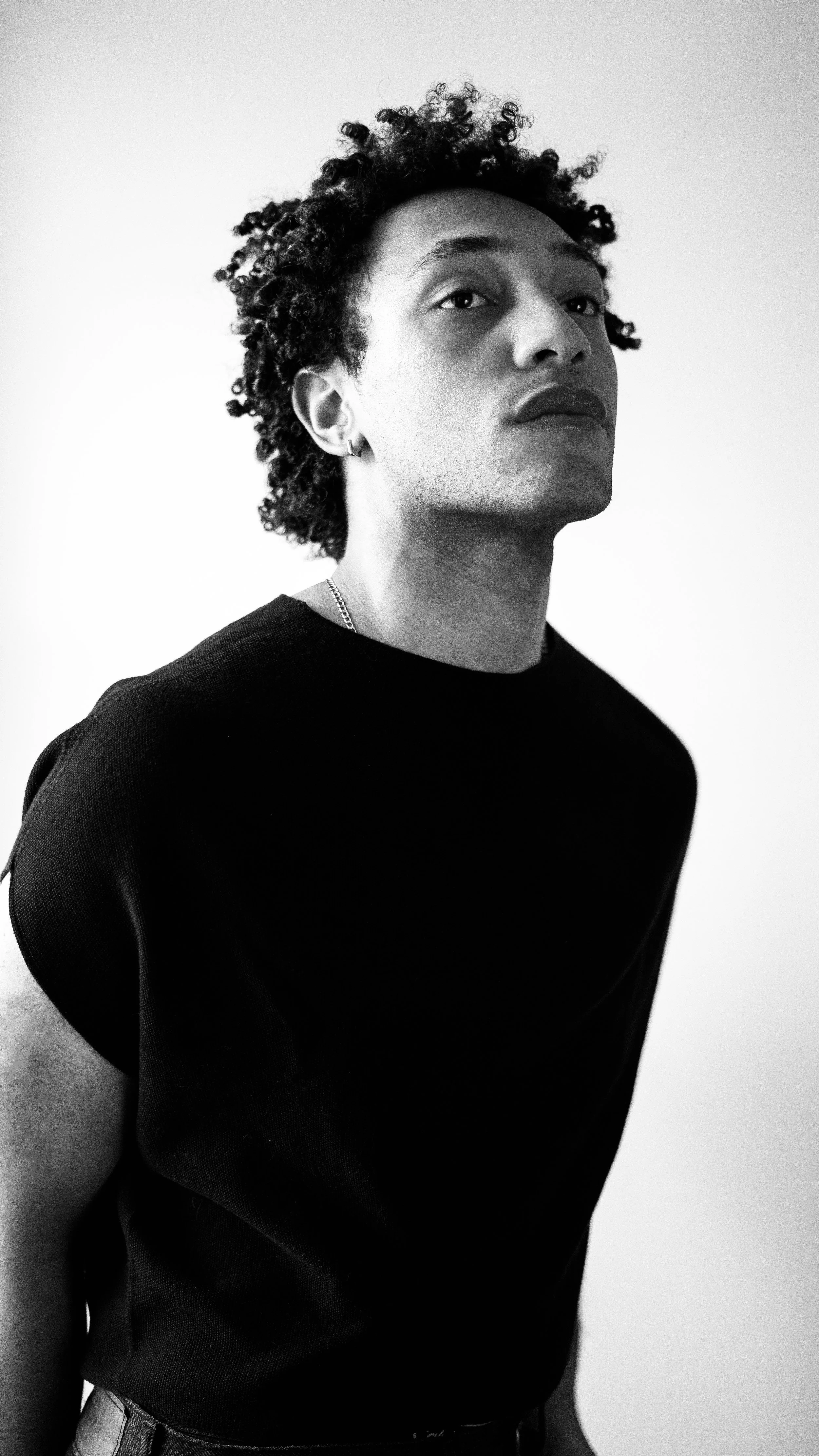 Black and white portrait of a young man with curly hair wearing a black shirt, looking directly at the camera with a neutral expression.