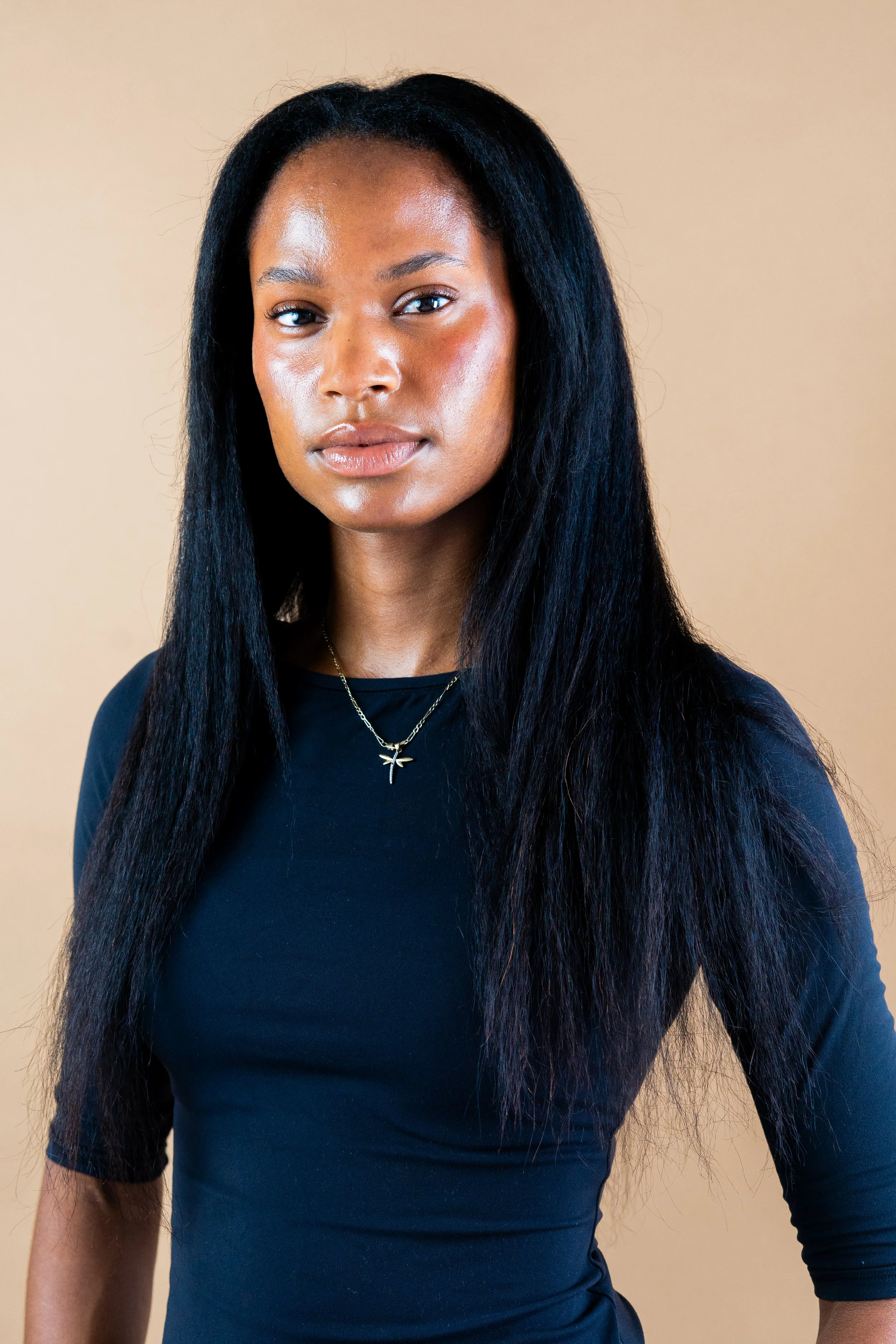 Close-up photo of a young woman with long black hair, wearing a black top and a silver dragonfly necklace, standing against a beige background.