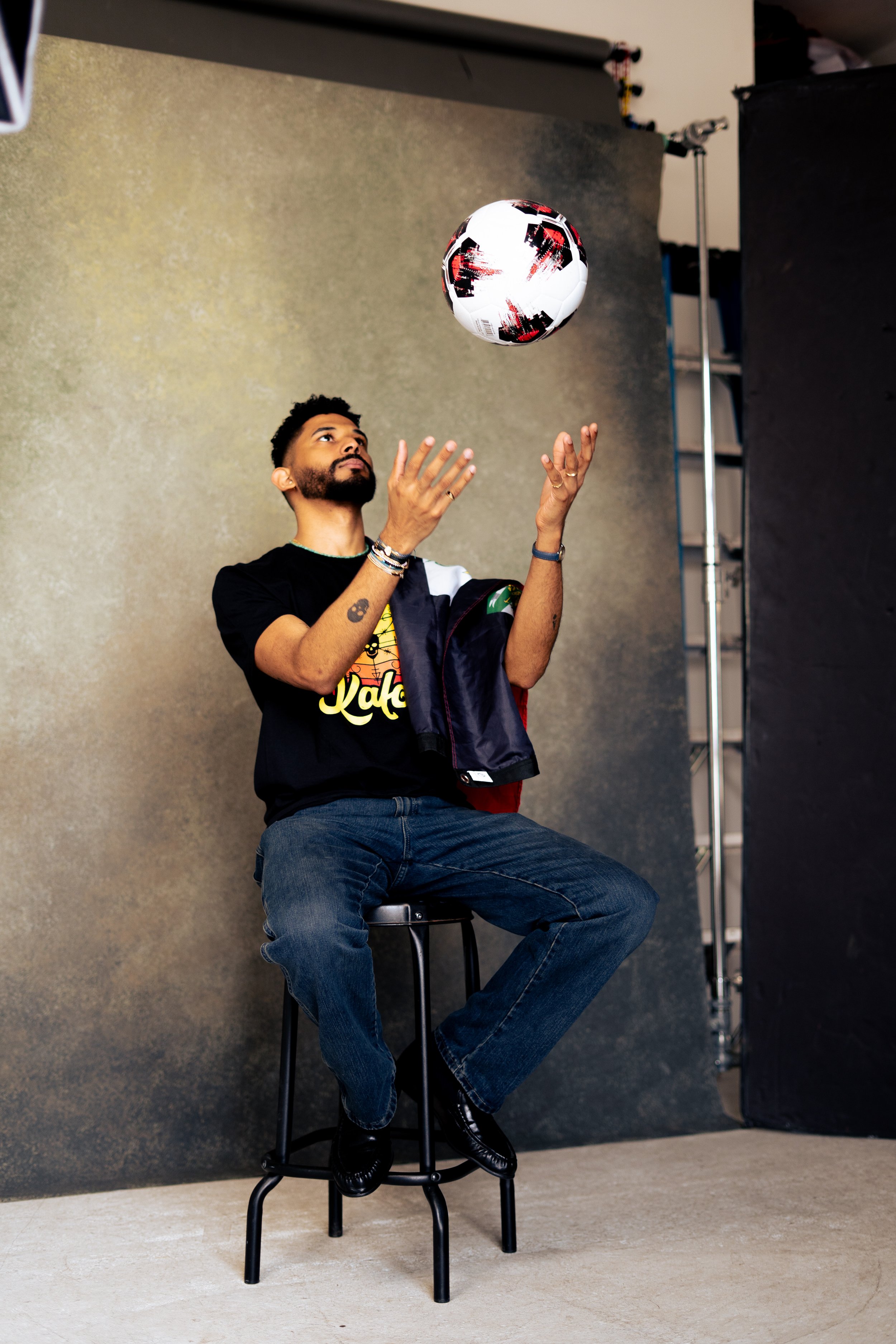 A man with dark curly hair and a beard is sitting on a tall black stool, wearing a black T-shirt with colorful text and graphics, jeans, and black shoes. He is indoors with a plain beige wall background and is playing with a black and white soccer ba