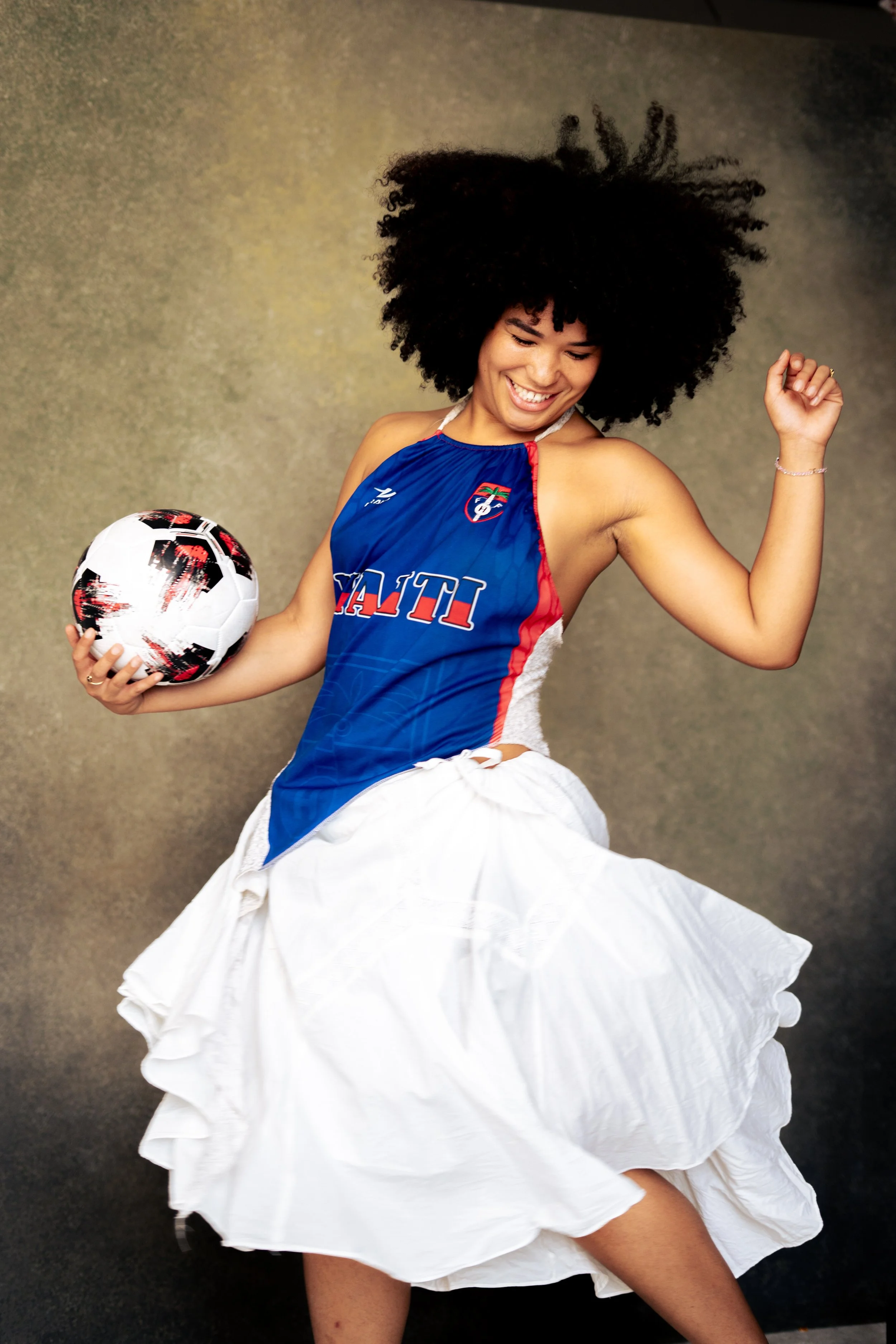 Woman smiling and dancing, holding a soccer ball, wearing a sports jersey and a white skirt, against a neutral background.