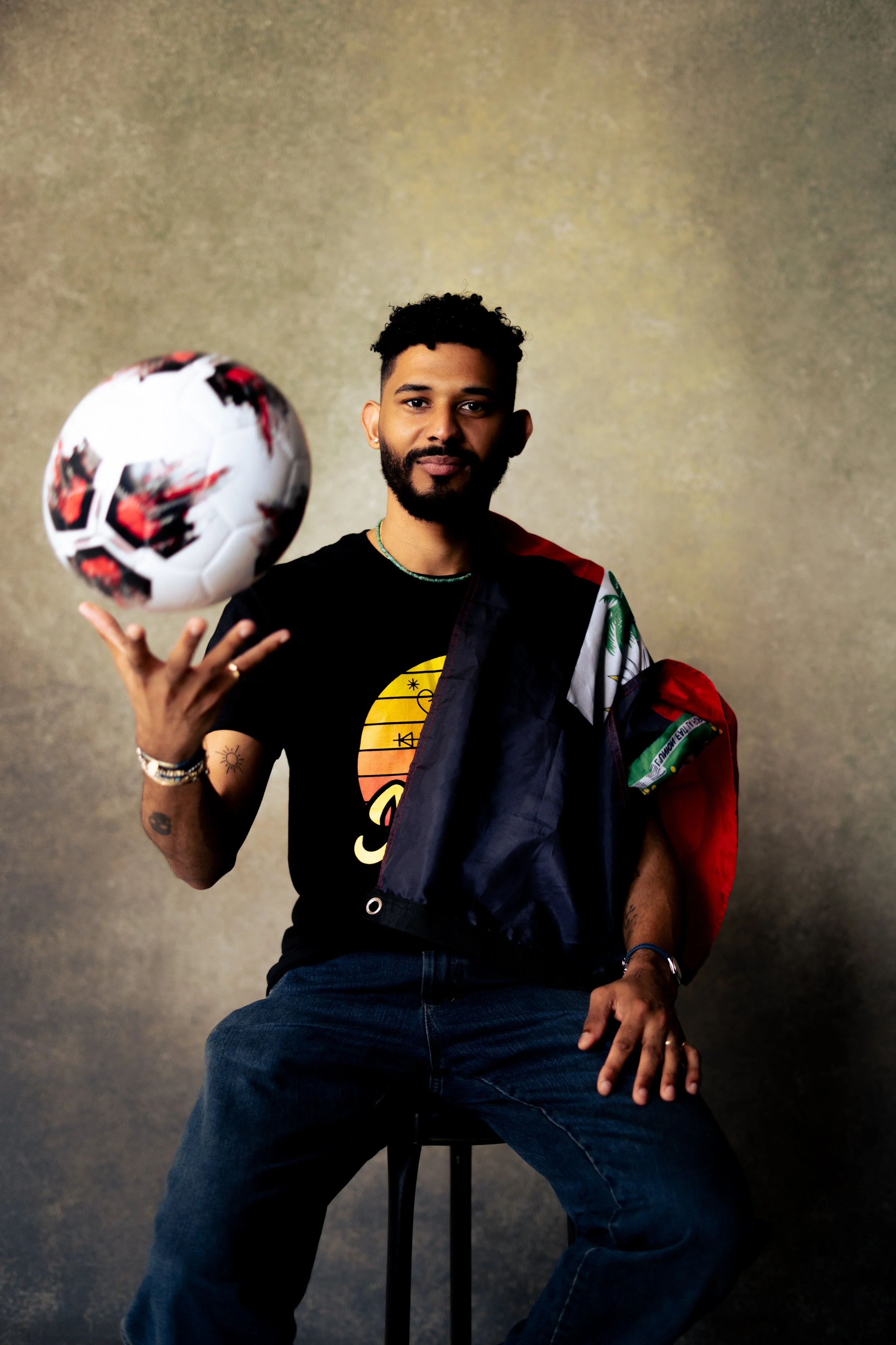 A man sitting on a black stool holding a soccer ball in his right hand, wearing a black t-shirt with a yellow and orange design and a rainbow flag draped over his shoulder, against a plain gray background.
