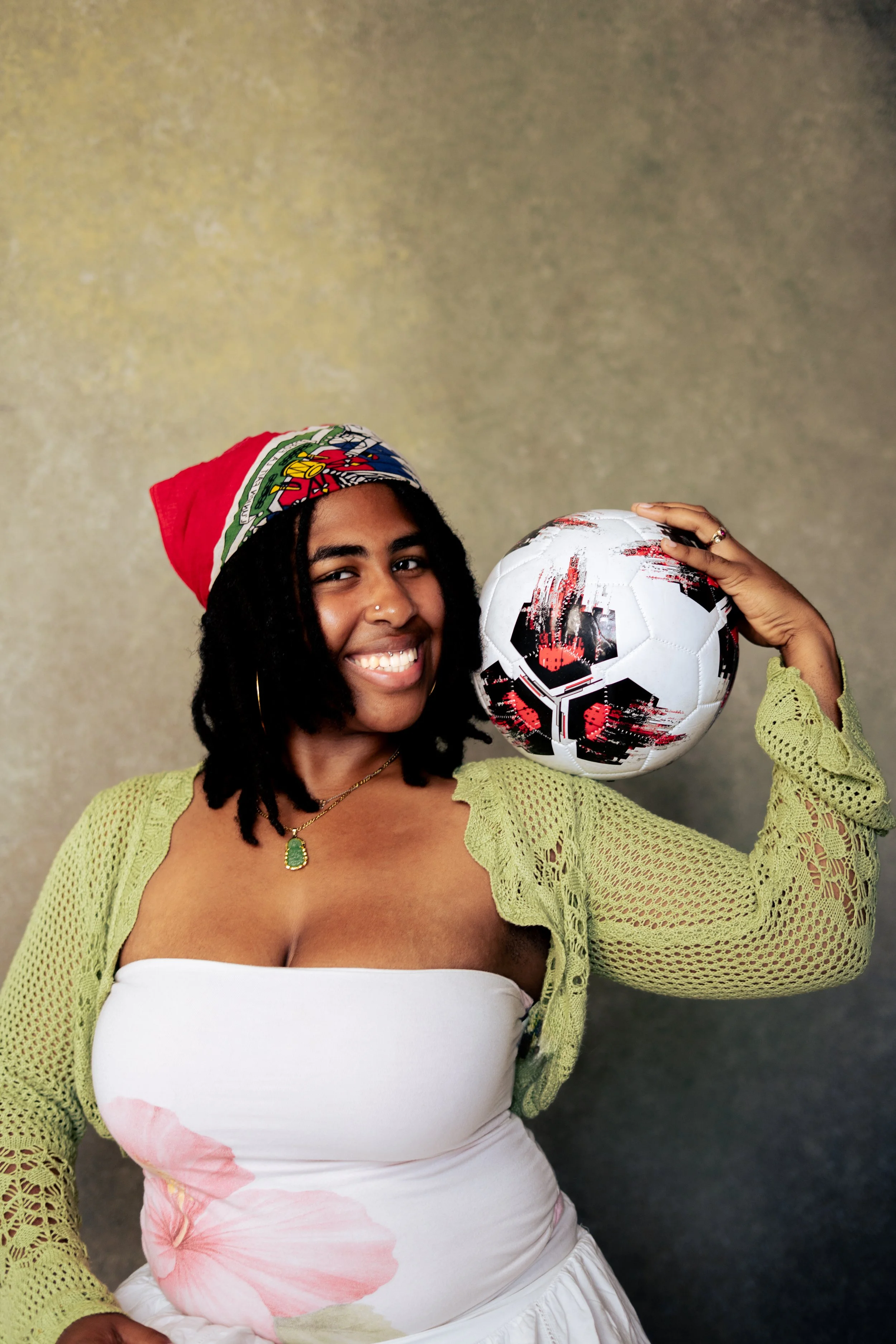 A smiling woman with dark curly hair wearing a green crochet top and a white strapless dress with pink floral prints, holding a black and white soccer ball, and wearing a red, white, and blue bandana, against a textured gray background.
