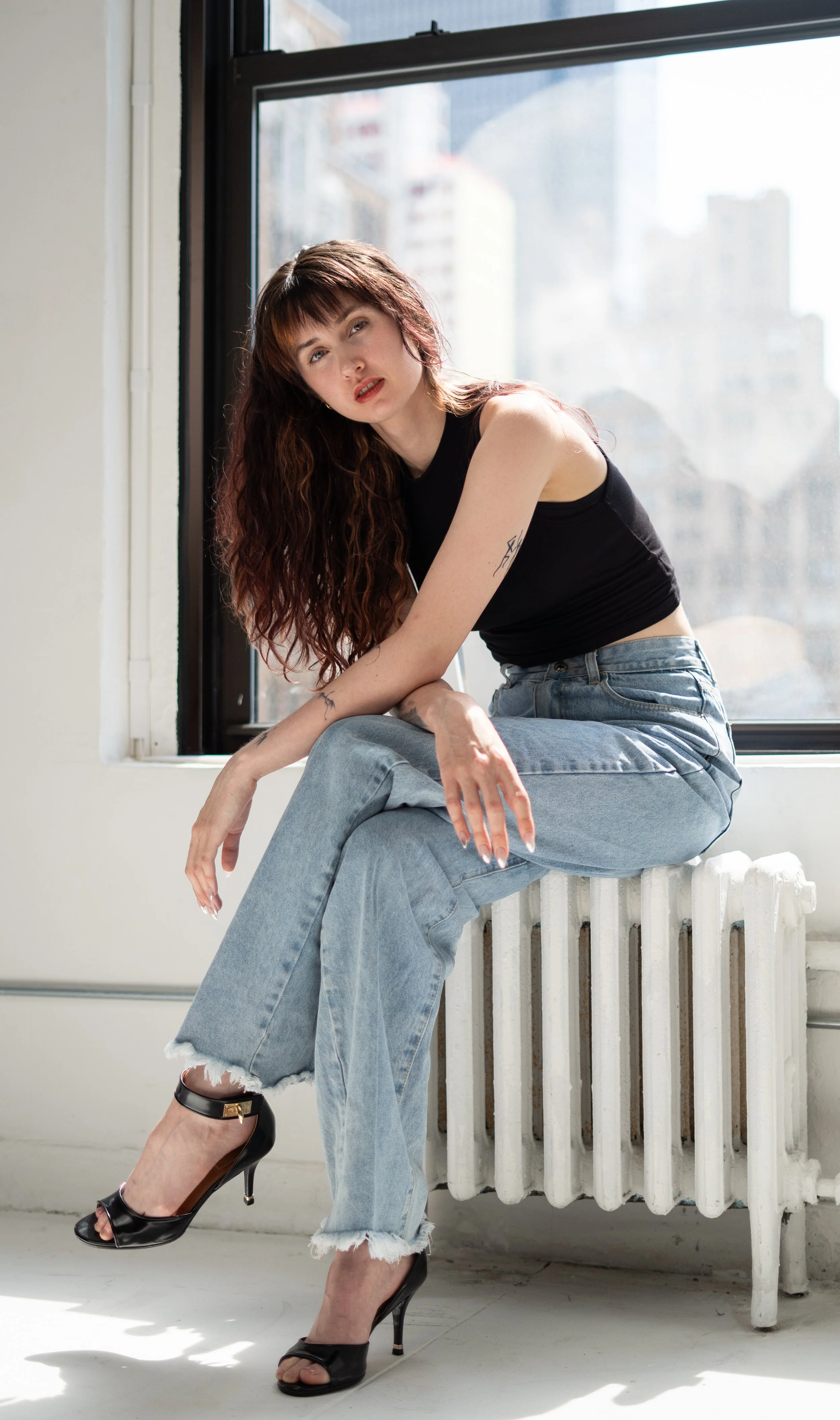 A young woman with long, wavy auburn hair sitting on a radiator by a window, wearing a black sleeveless top, light blue ripped jeans, and black high heels, looking at the camera with a relaxed expression.