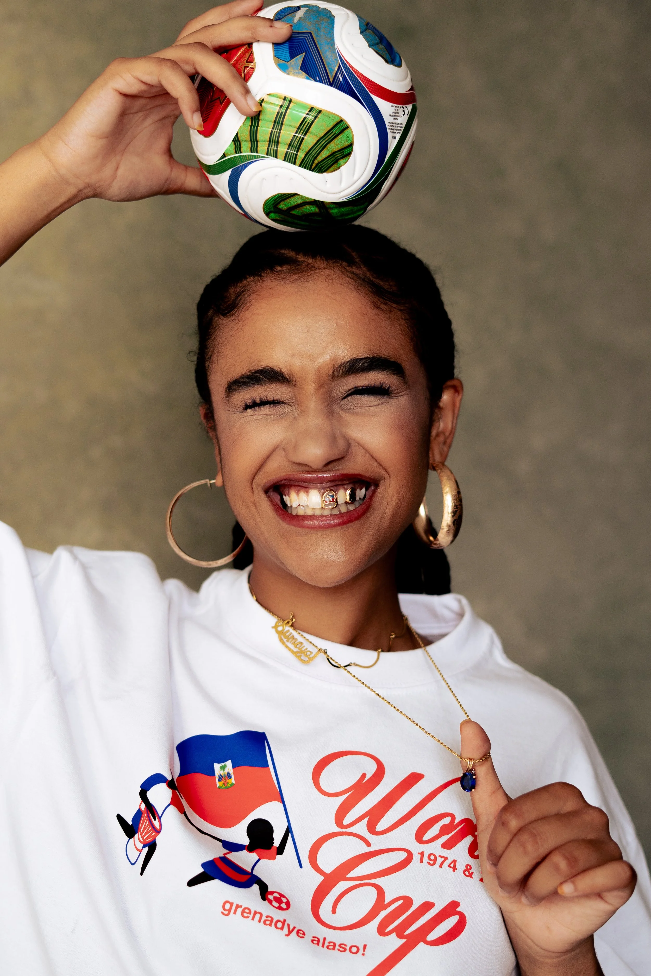A young woman with curly hair and large hoop earrings is holding a colorful soccer ball on her head. She is smiling with her teeth showing, wearing gold jewelry including a necklace with a nameplate, and a white t-shirt with an illustration and text 