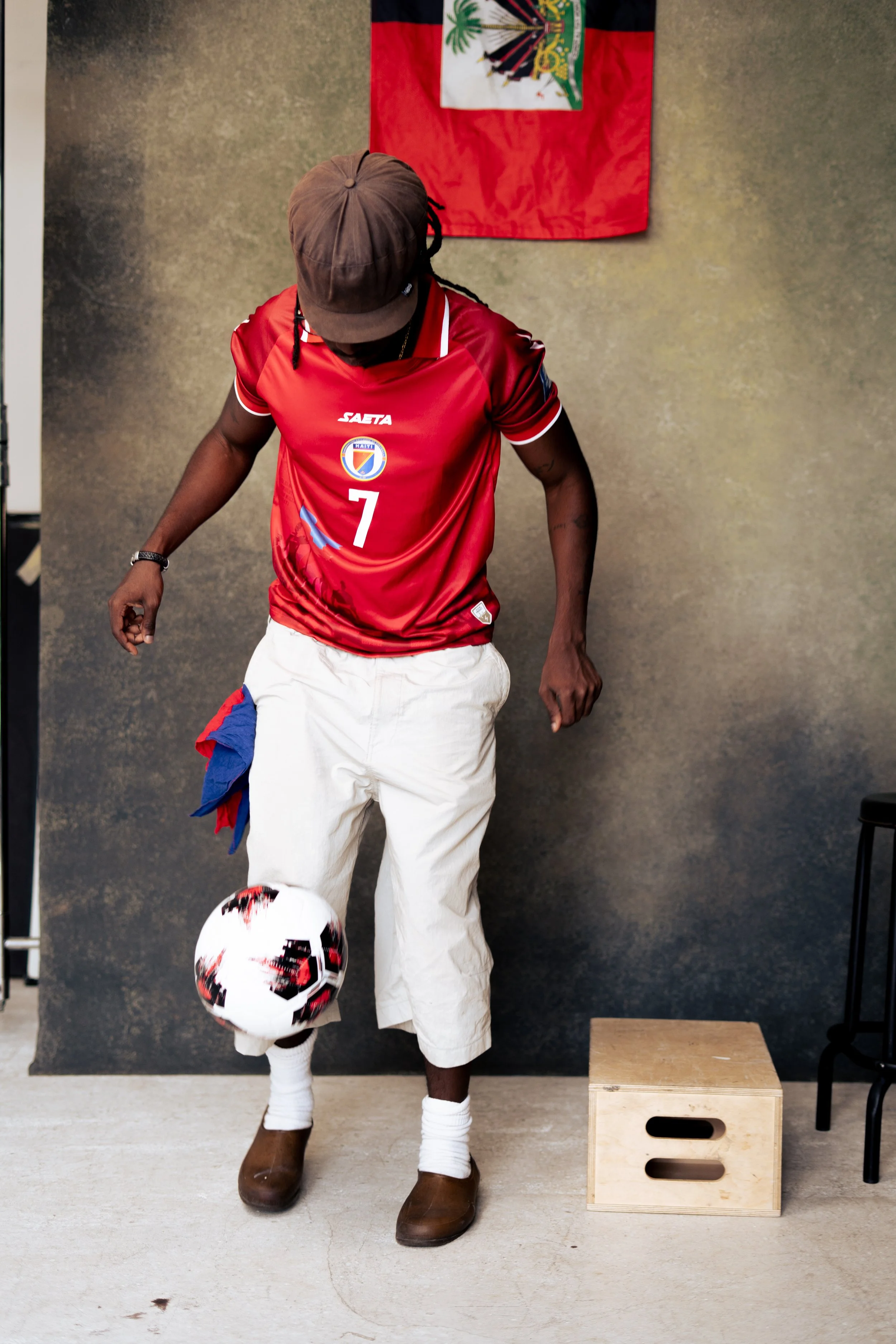 A man wearing a brown cap, red soccer jersey with the number 7, white pants, and brown shoes, is standing and playing with a soccer ball in front of a gray wall with a Haitian flag hanging on it.