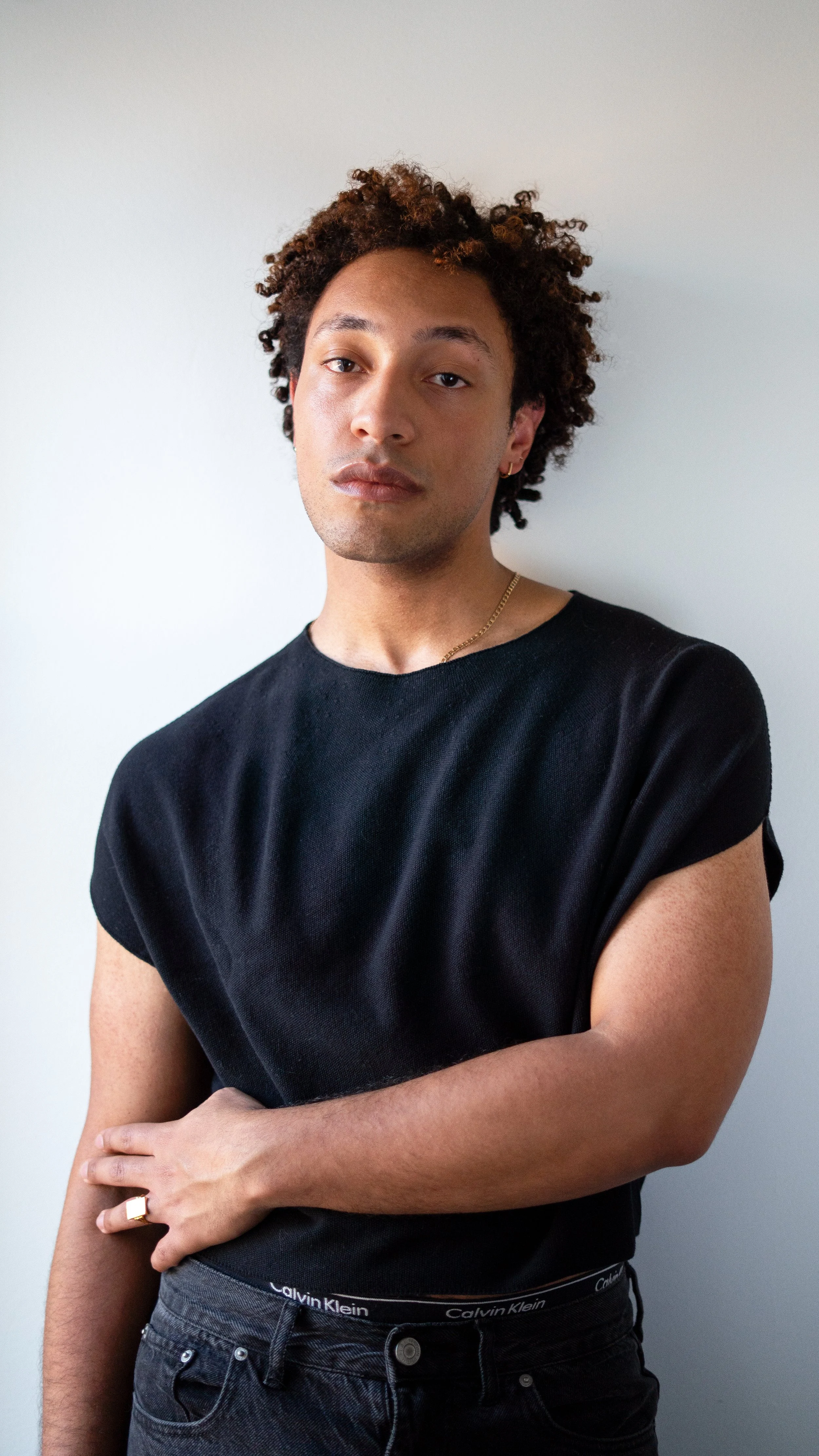 A young person standing against a gray wall, wearing a black t-shirt, dark jeans, and a gold chain, with short, curly hair and a serious expression.