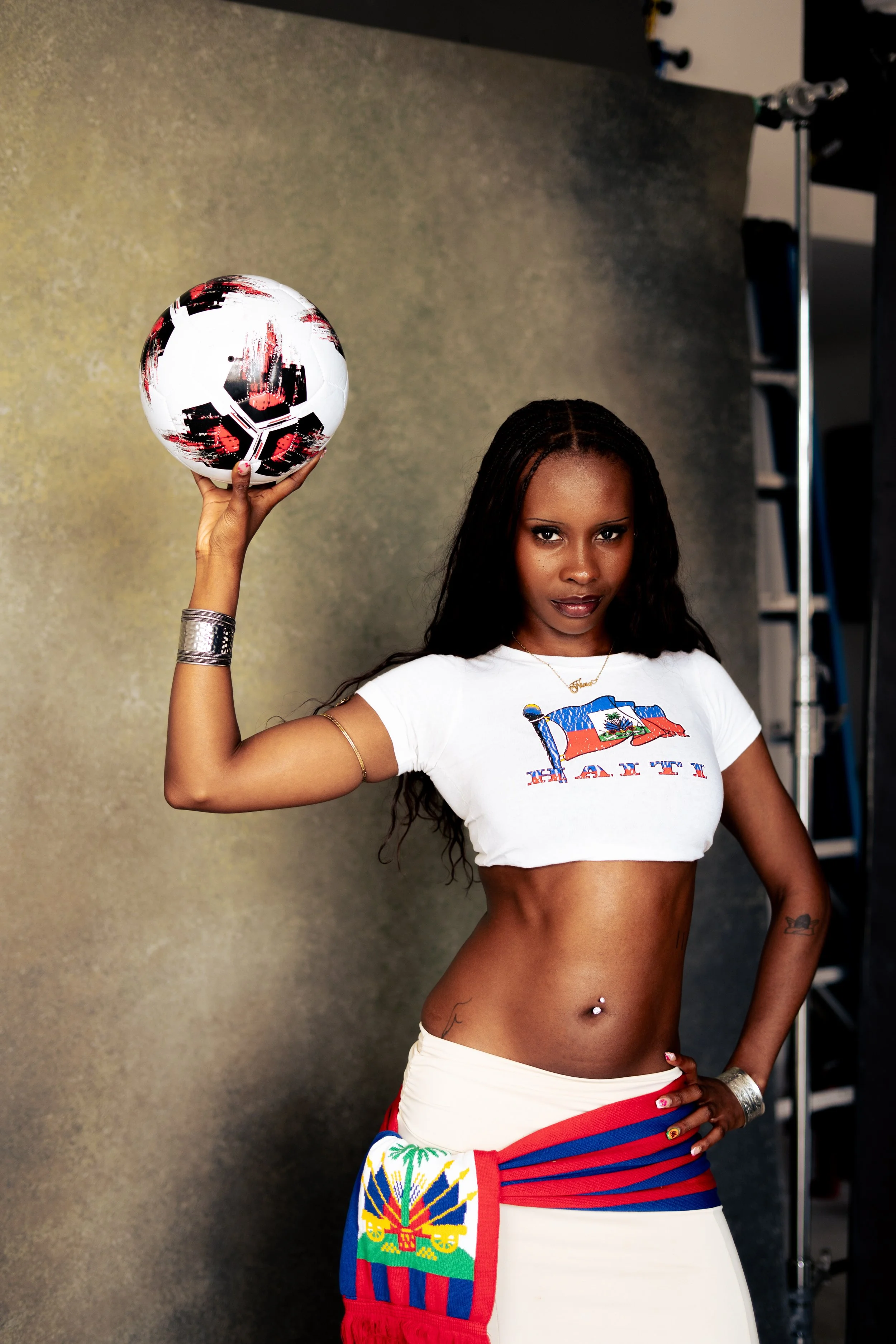 A woman with dark hair and medium skin tone holding a black, white, and red soccer ball above her head. She is wearing a cropped T-shirt with the Haiti flag and a white skirt with colorful patterns. She has tattoos, jewelry, and a confident expressio