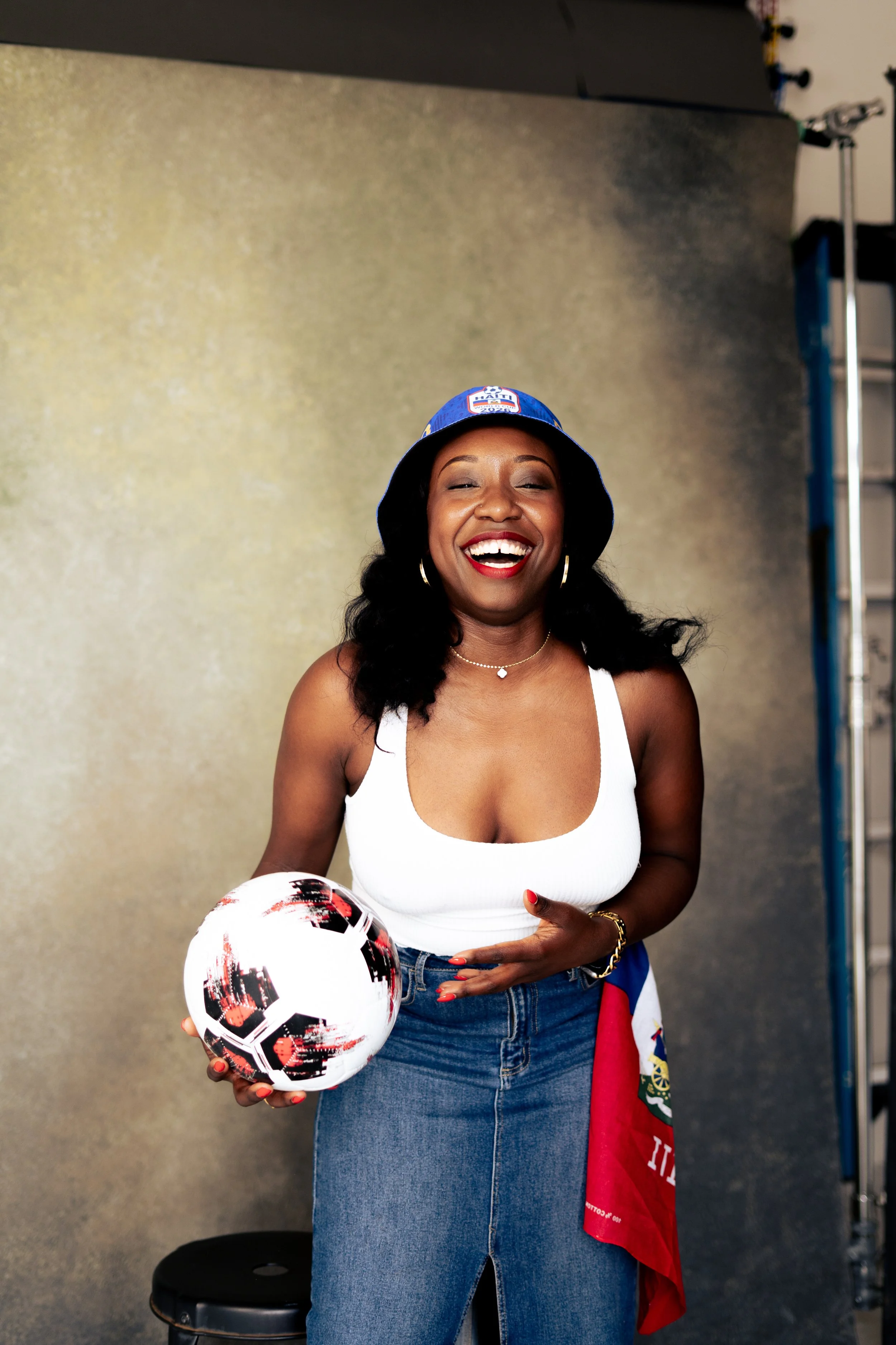 A woman with dark hair, wearing a blue bucket hat, a white tank top, and jeans, smiling and holding a soccer ball in her left hand. She is standing in front of a beige wall, with a flag and equipment in the background.