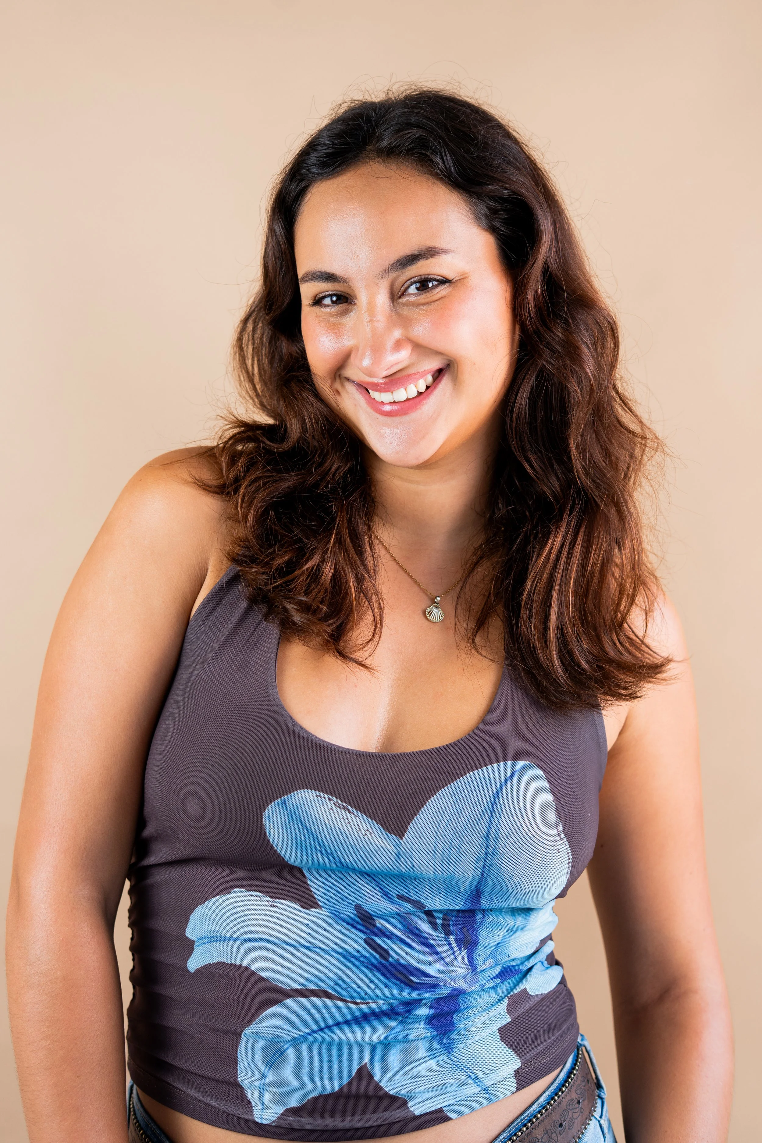 Portrait of a woman with wavy brown hair, smiling, wearing a gray tank top with a large blue flower print and light blue jeans with a studded belt, standing against a plain beige background.