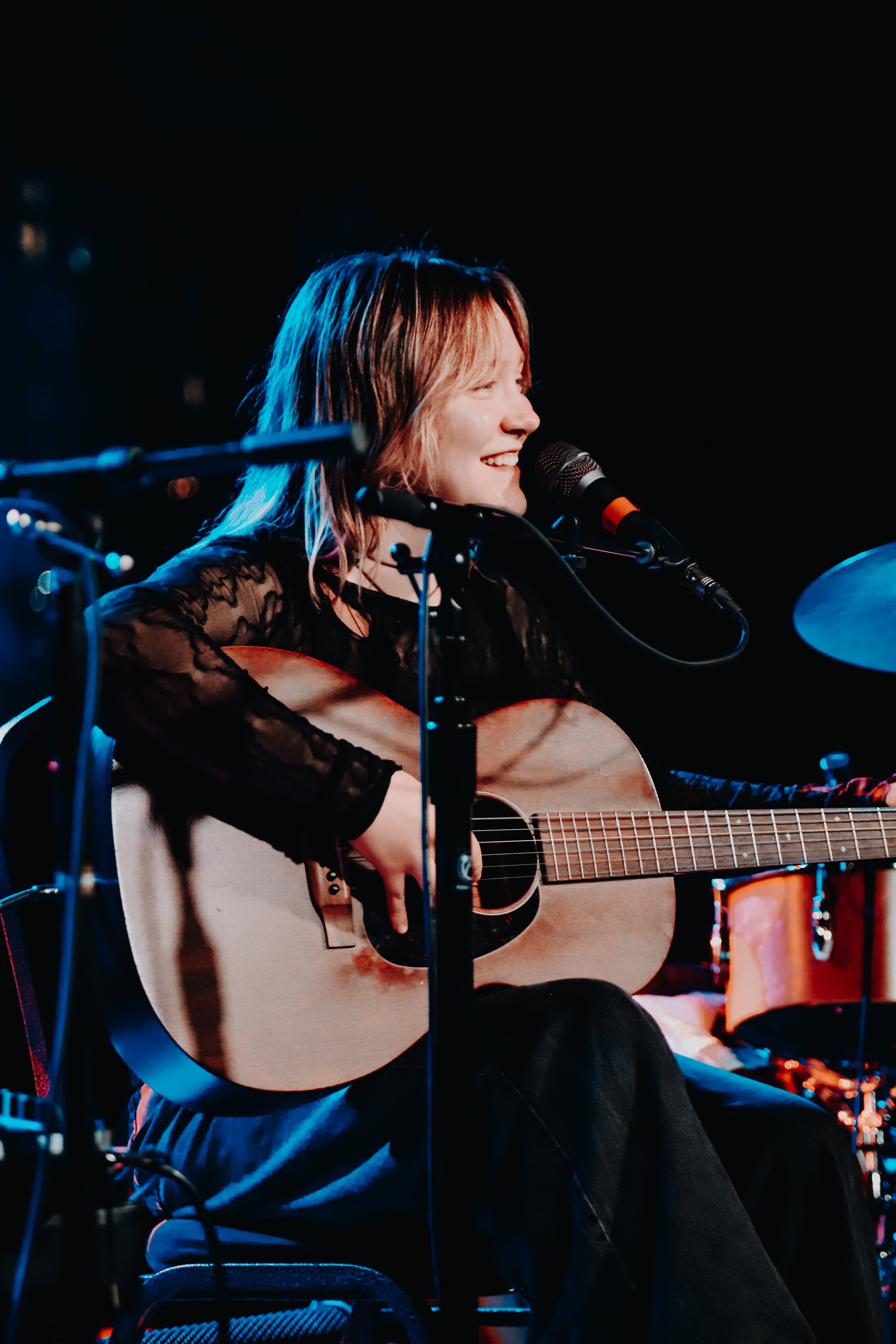 A woman with shoulder-length hair, smiling and playing an acoustic guitar, singing into a microphone during a live performance in a dimly lit setting.