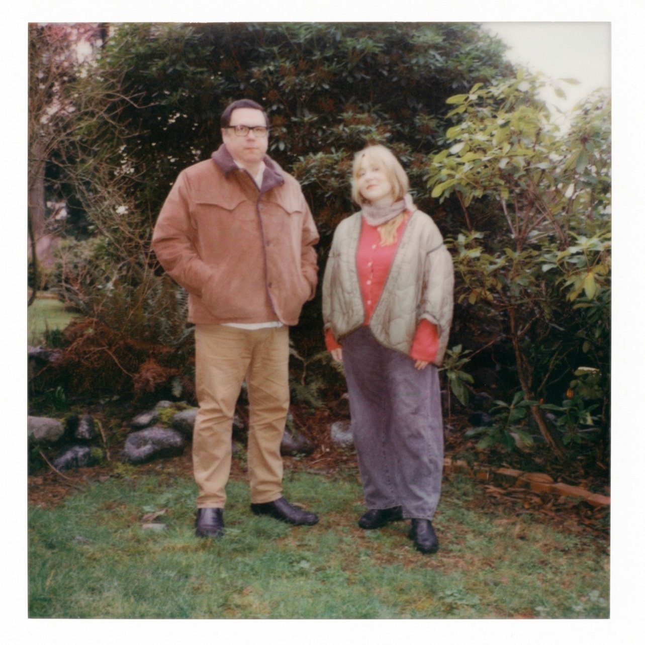 A man and woman standing outdoors in front of bushes and trees, dressed in warm clothing, with the man having short dark hair and glasses, and the woman with shoulder-length blonde hair.