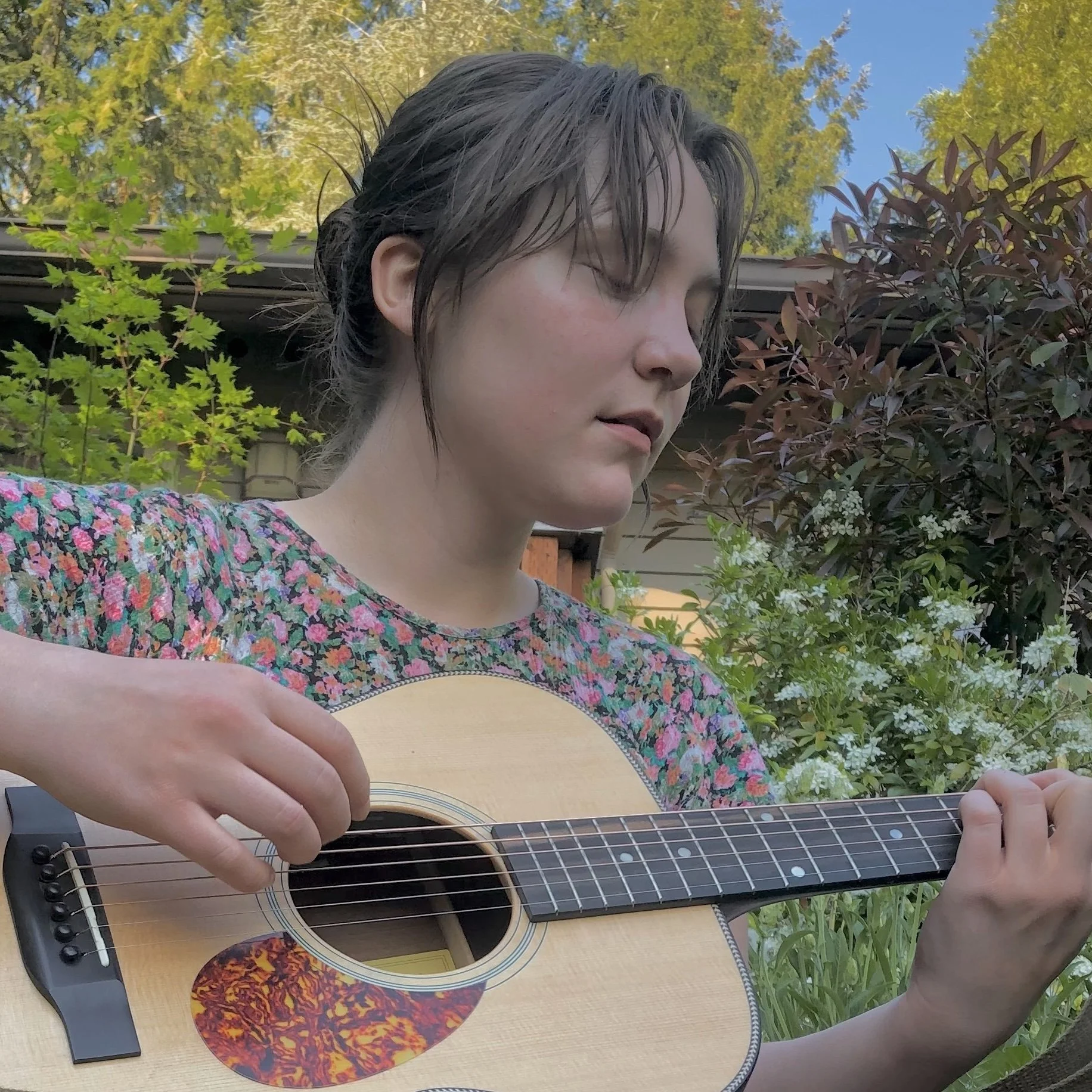 A young woman with short dark hair playing an acoustic guitar outdoors, surrounded by green plants and trees under a clear blue sky.
