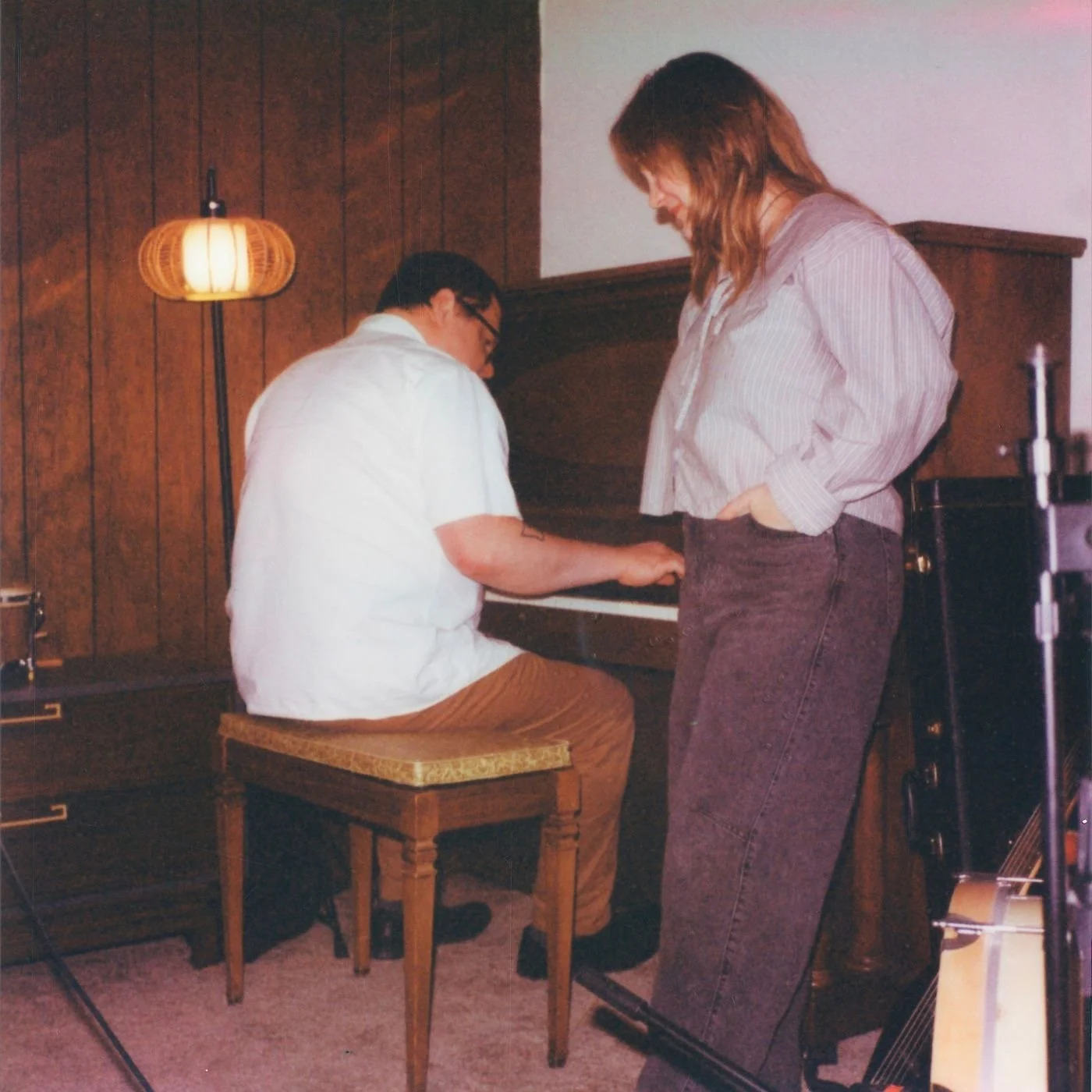 A man sitting on a wooden stool playing the piano while a woman standing beside him watches, in a room with wood-paneled walls and a lamp hanging from the ceiling.