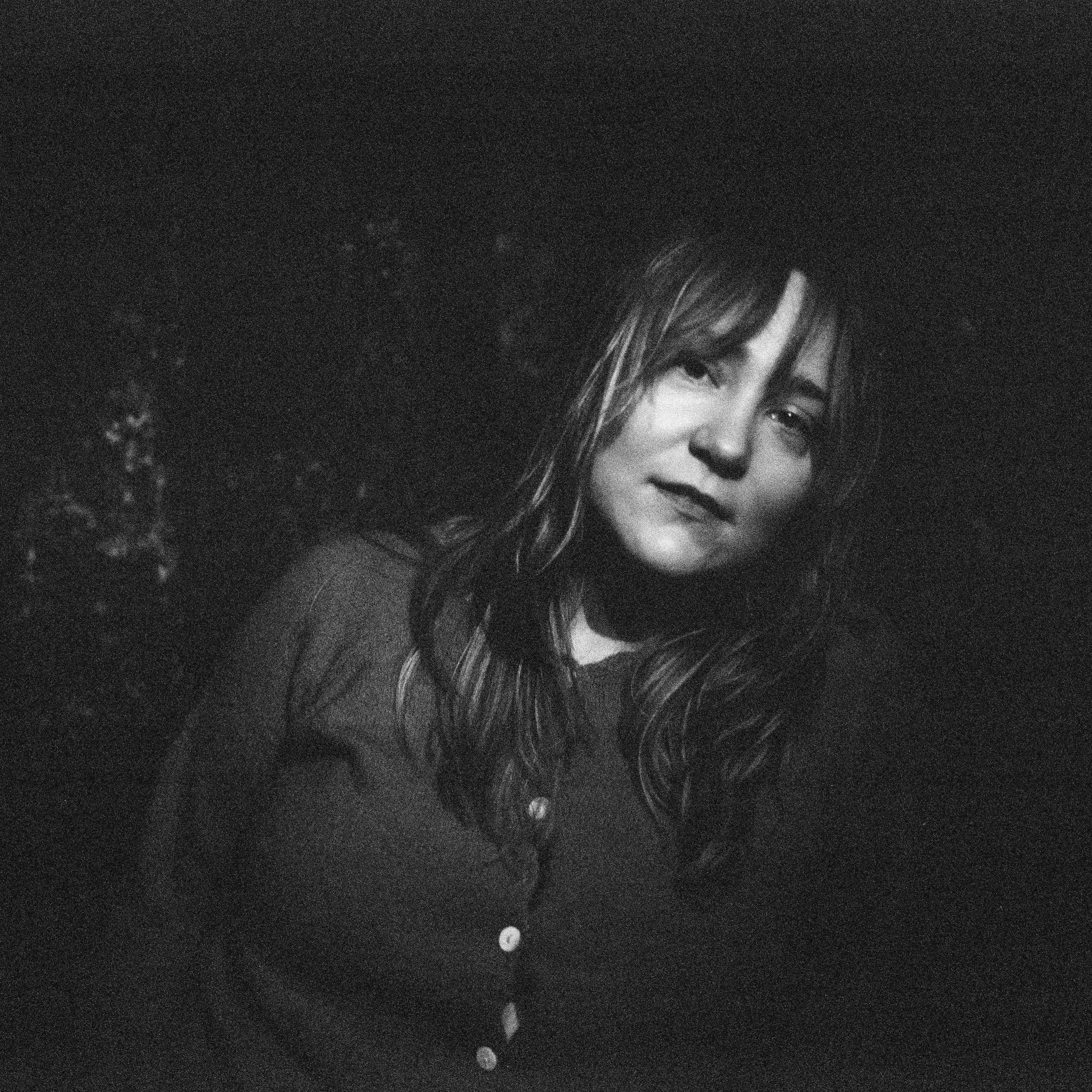 A young woman with wavy hair looking at the camera, with a slight smile, in a dark setting in black and white.