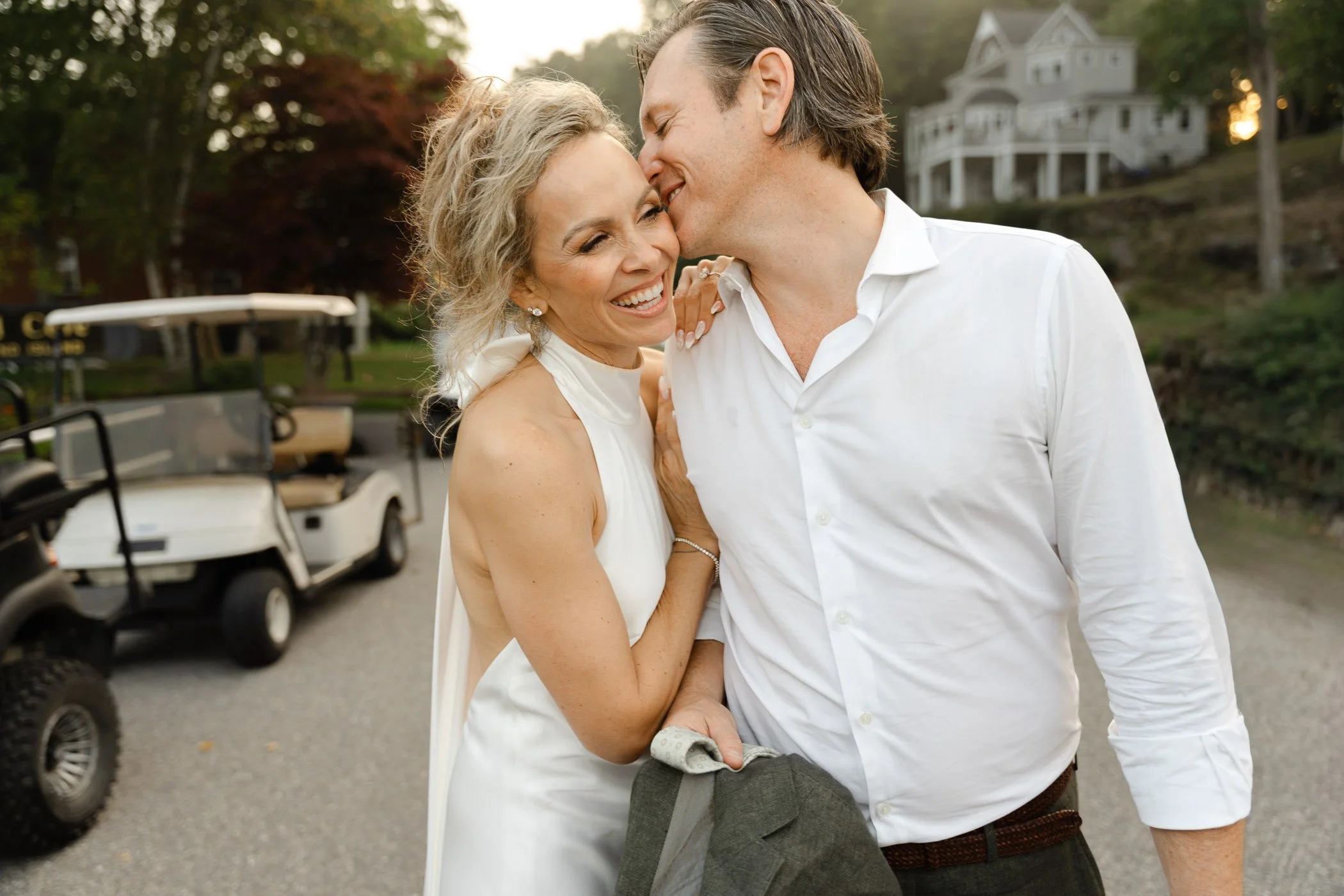 A smiling couple embraces on a road, with the man kissing the womans cheek; a golf cart and house are in the background.