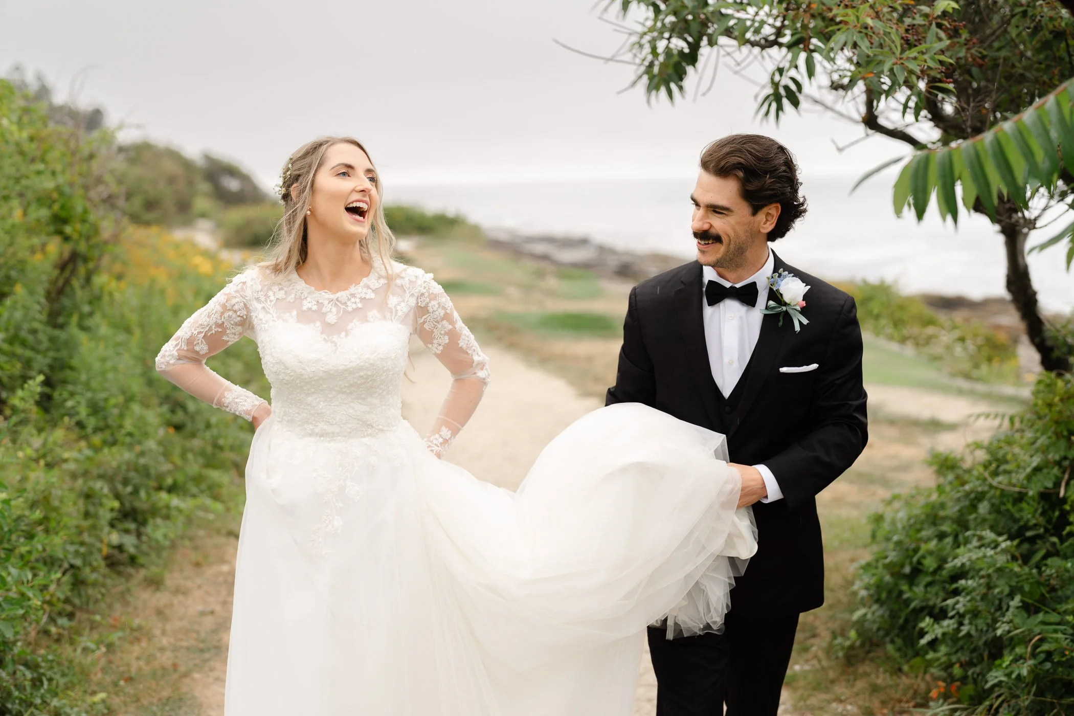 Bride laughing as groom in tuxedo holds her dress train at their Maine wedding, walking down a path surrounded by greenery.