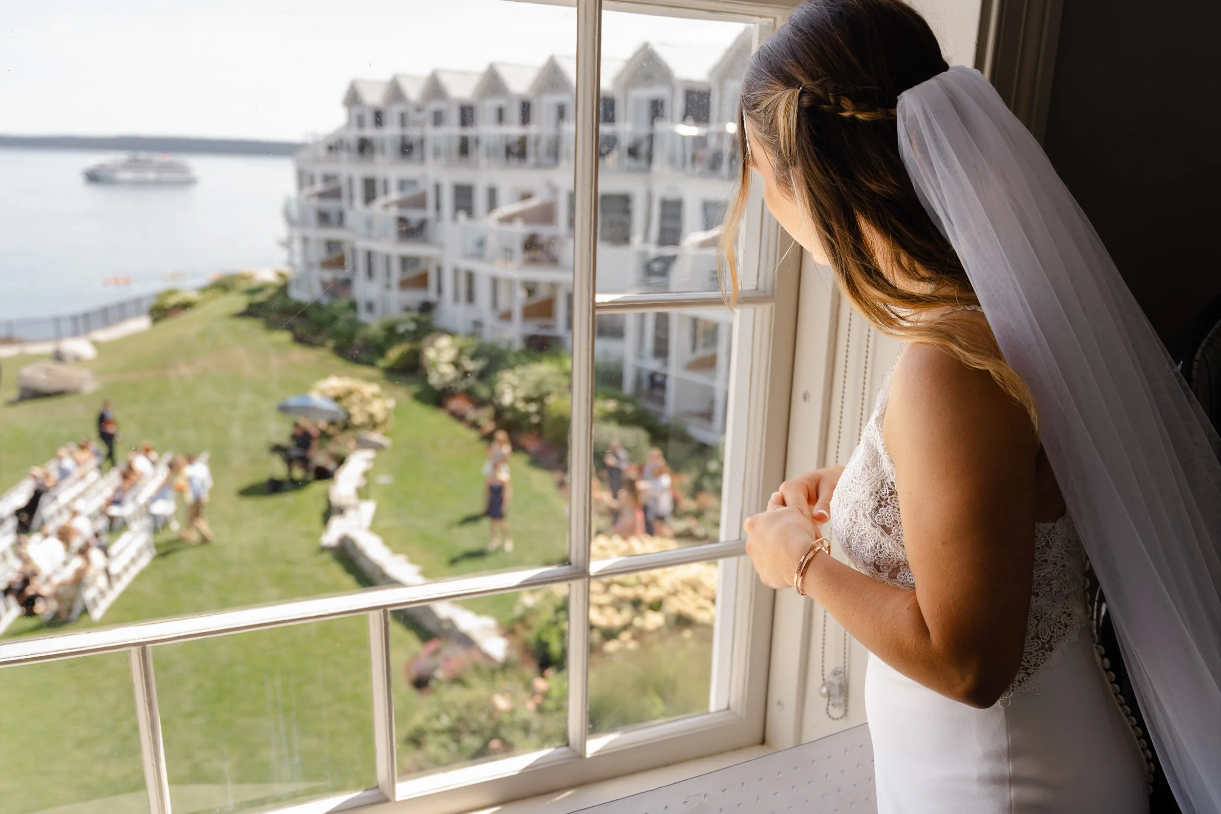 A bride in a white dress and veil looks out a window at a waterfront hotel wedding setup on a sunny day.