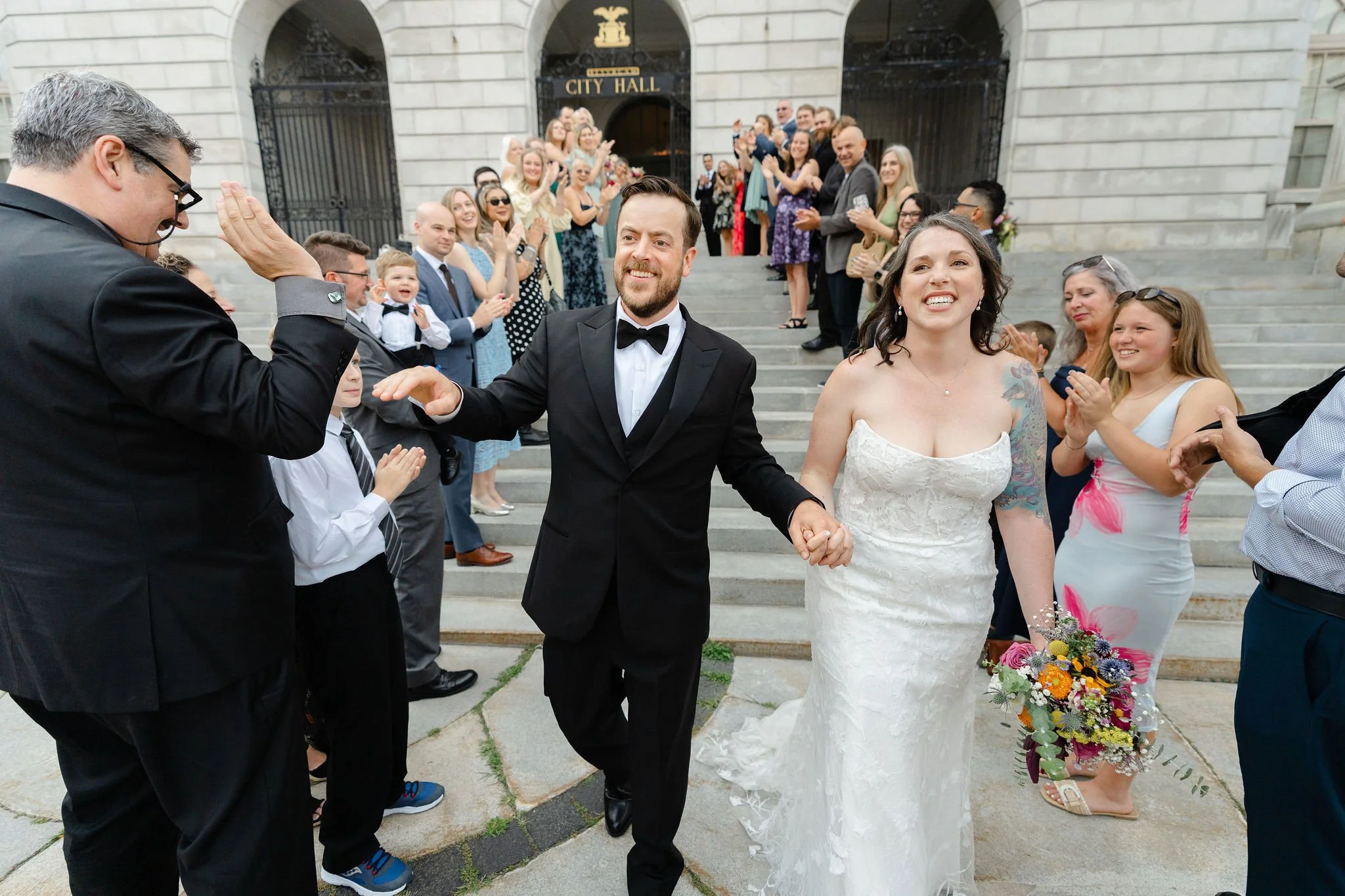 A bride and groom walk hand in hand outside city hall as guests cheer and celebrate their wedding.