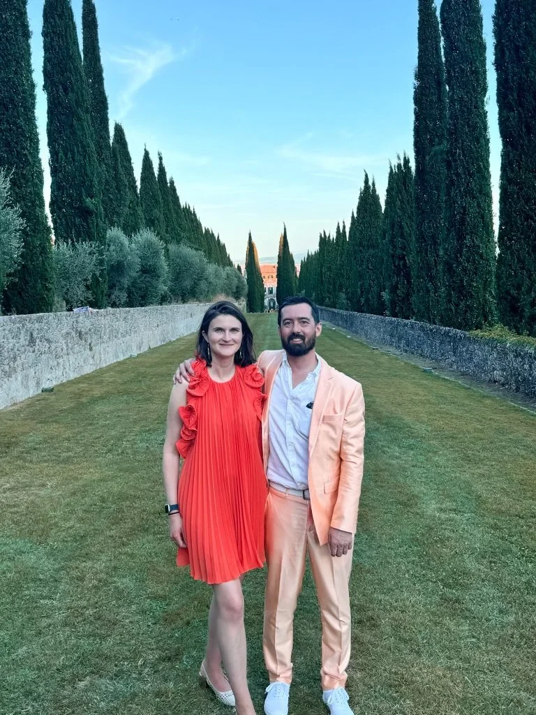 A man in a tuxedo and a woman in a patterned dress smile on a city rooftop with tall buildings behind them. Ned and Julia Warner at a wedding in Italy