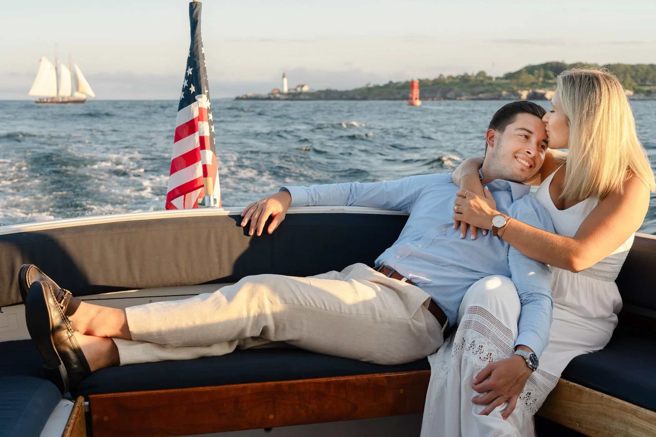A couple in white outfits cuddles on a boat, with an American flag, ocean waves, and a lighthouse in the background.