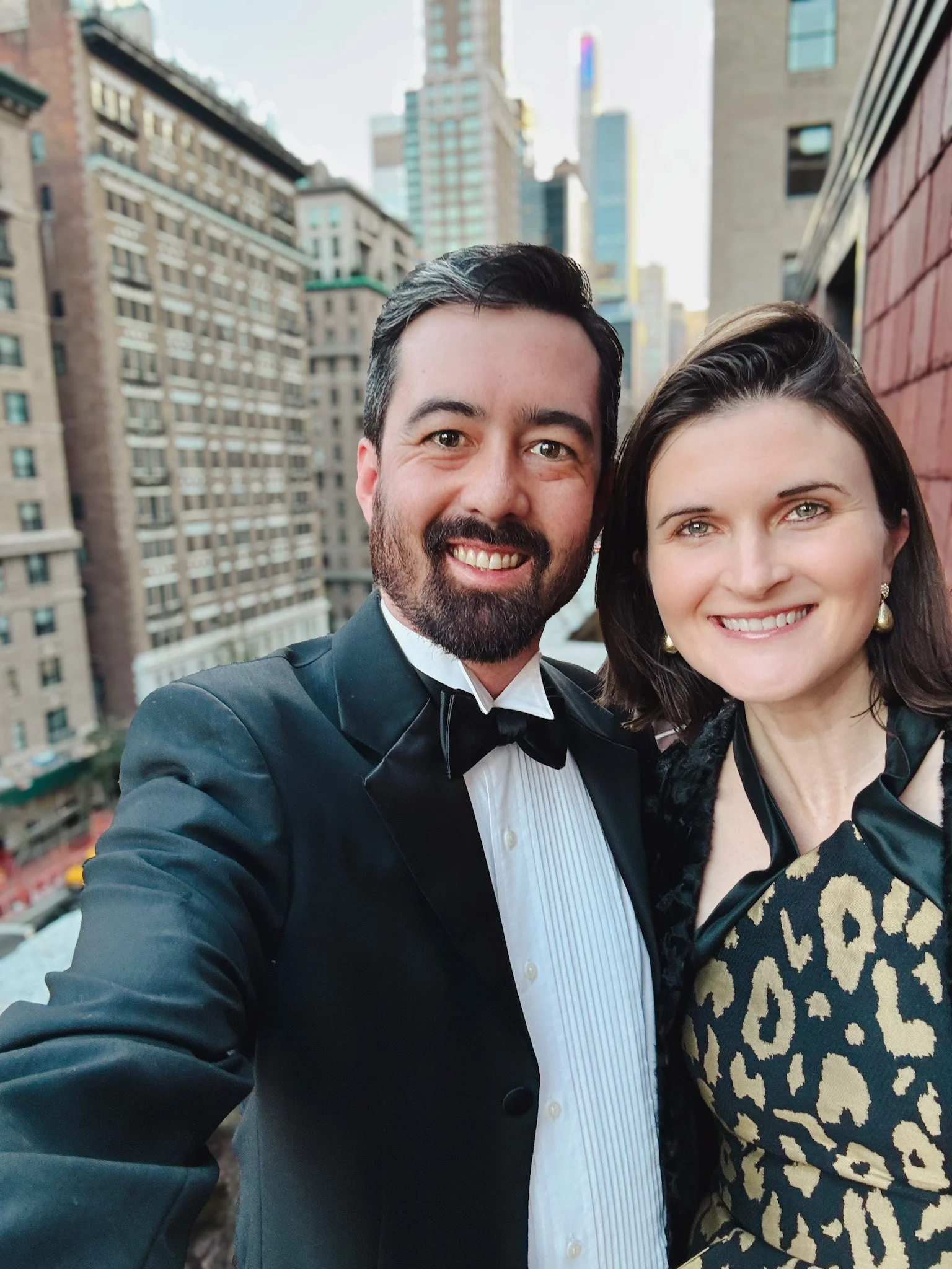A man in a tuxedo and a woman in a patterned dress smile on a city rooftop with tall buildings behind them. Ned and Julia Warner