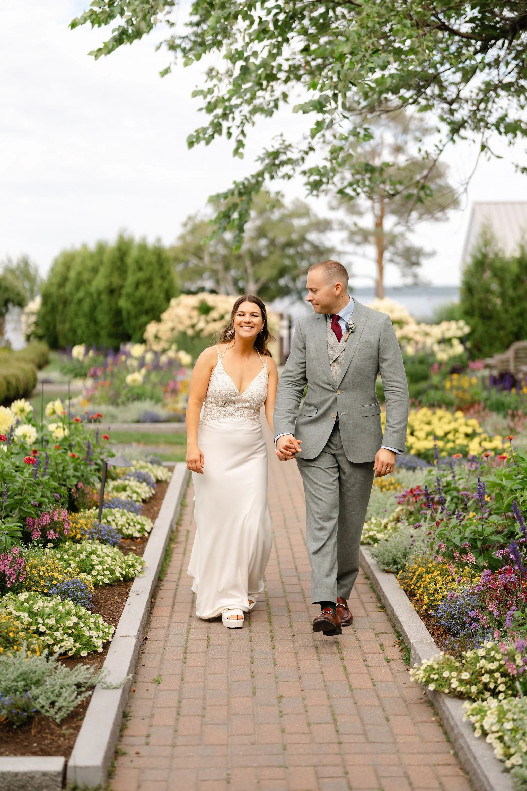 A happy couple dressed in formal wedding attire, standing outdoors in a wooded area. The man is wearing a dark blue tuxedo with a black bow tie. captured by Maine wedding photographers Warner Photography at Migis Lodge