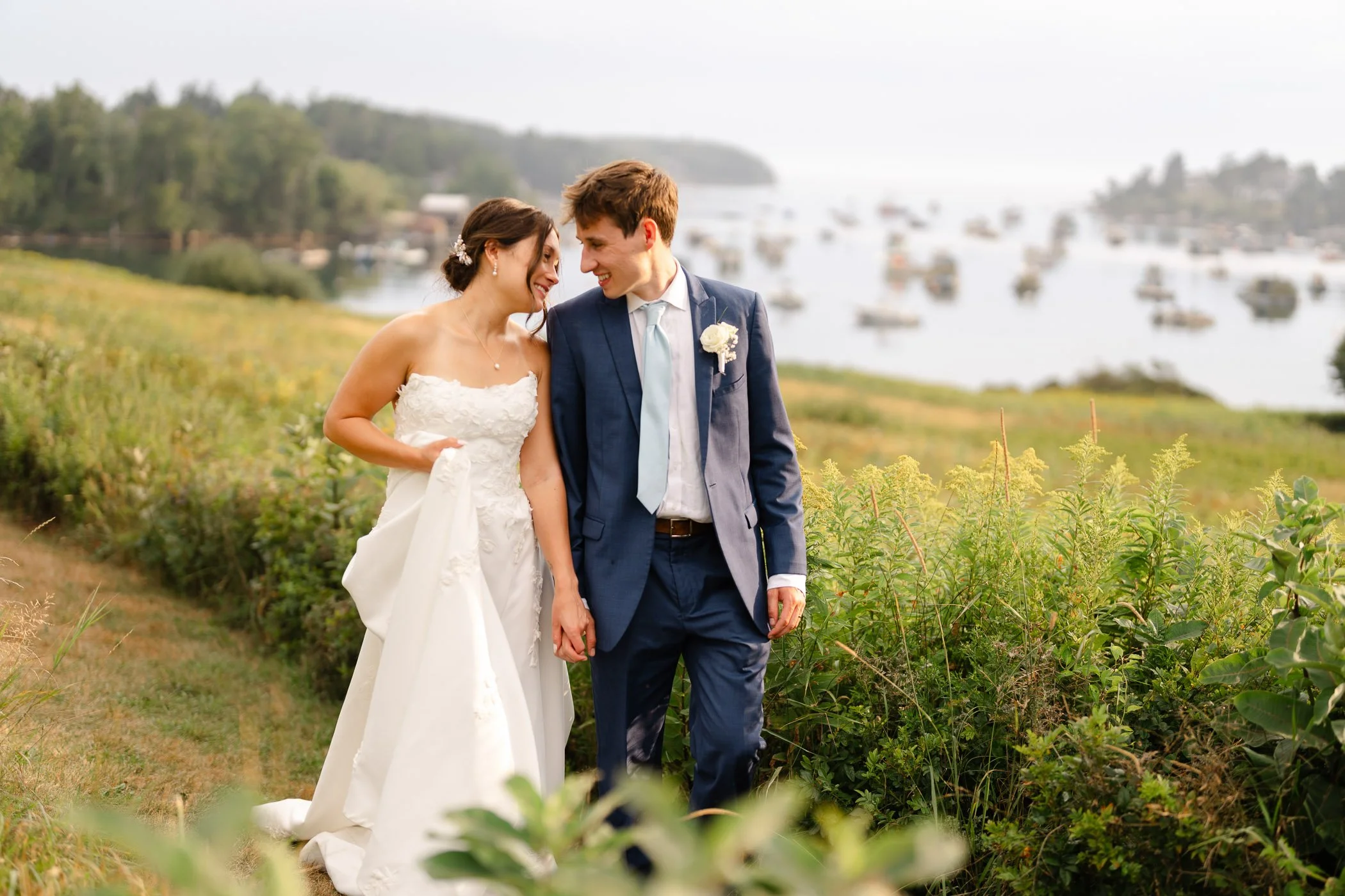 Bride and groom in wedding attire smile at each other, walking outdoors with water and boats in the background.
