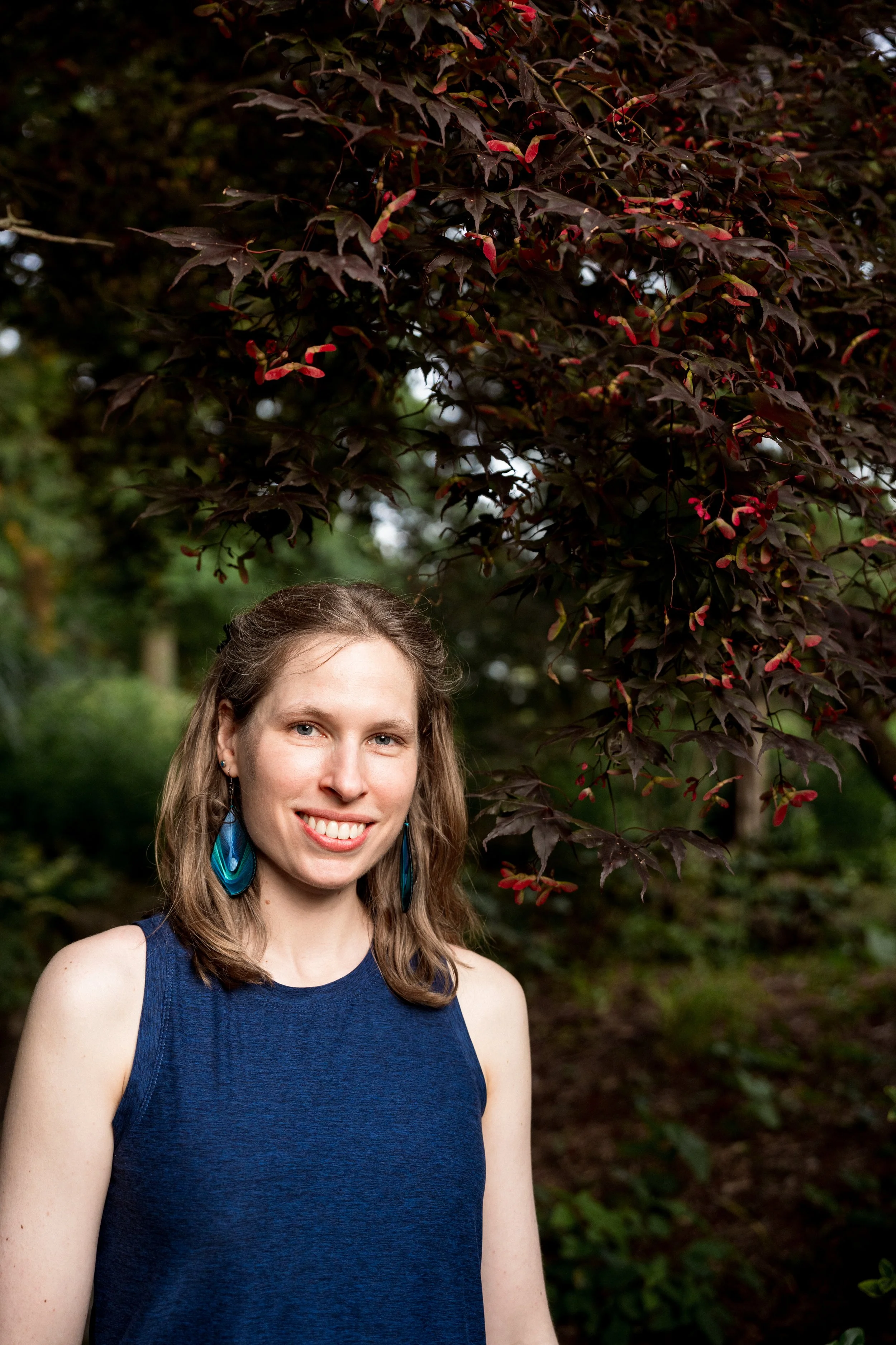 Anna, founder of Take Heart Yoga Therapy, smiling warmly outdoors under a tree. She is shown from the waist up.