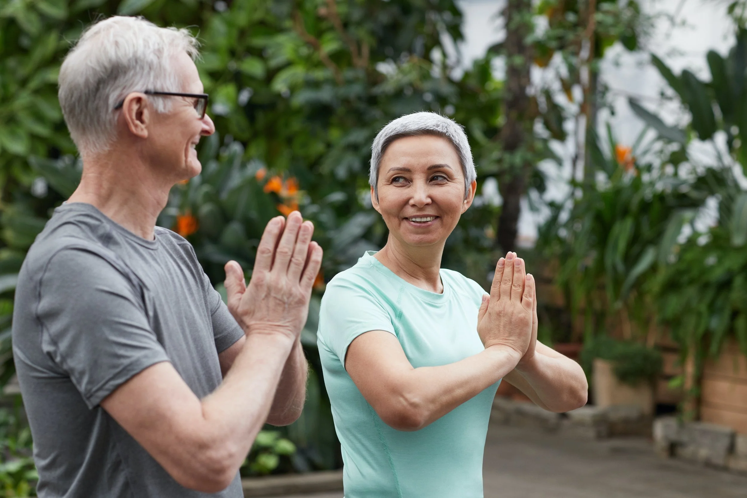 Two older adults standing side by side with hands at heart centre with palms touching  looking at one another and smiling.