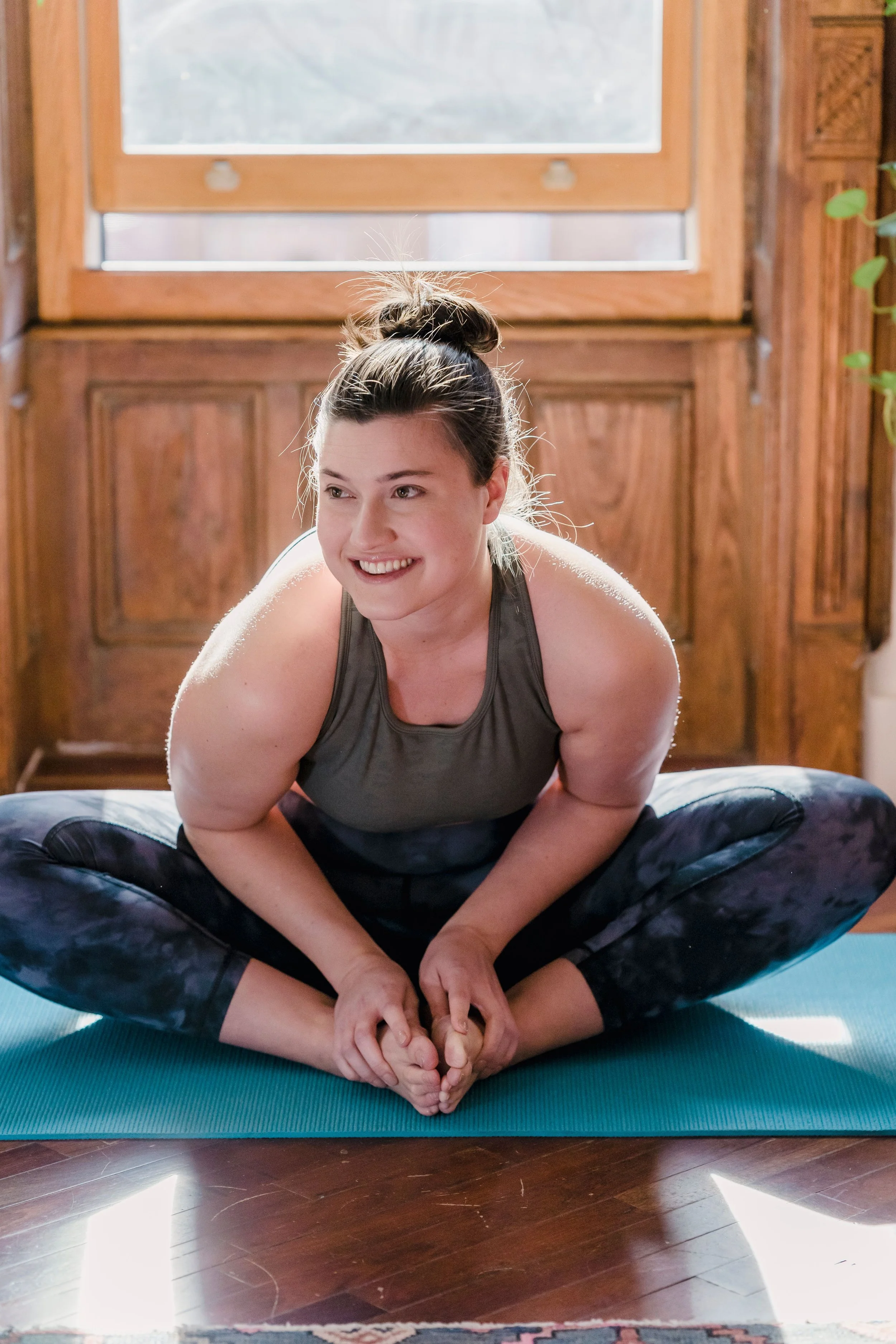 A woman smiling in butterfly pose, sunlit on her yoga mat.