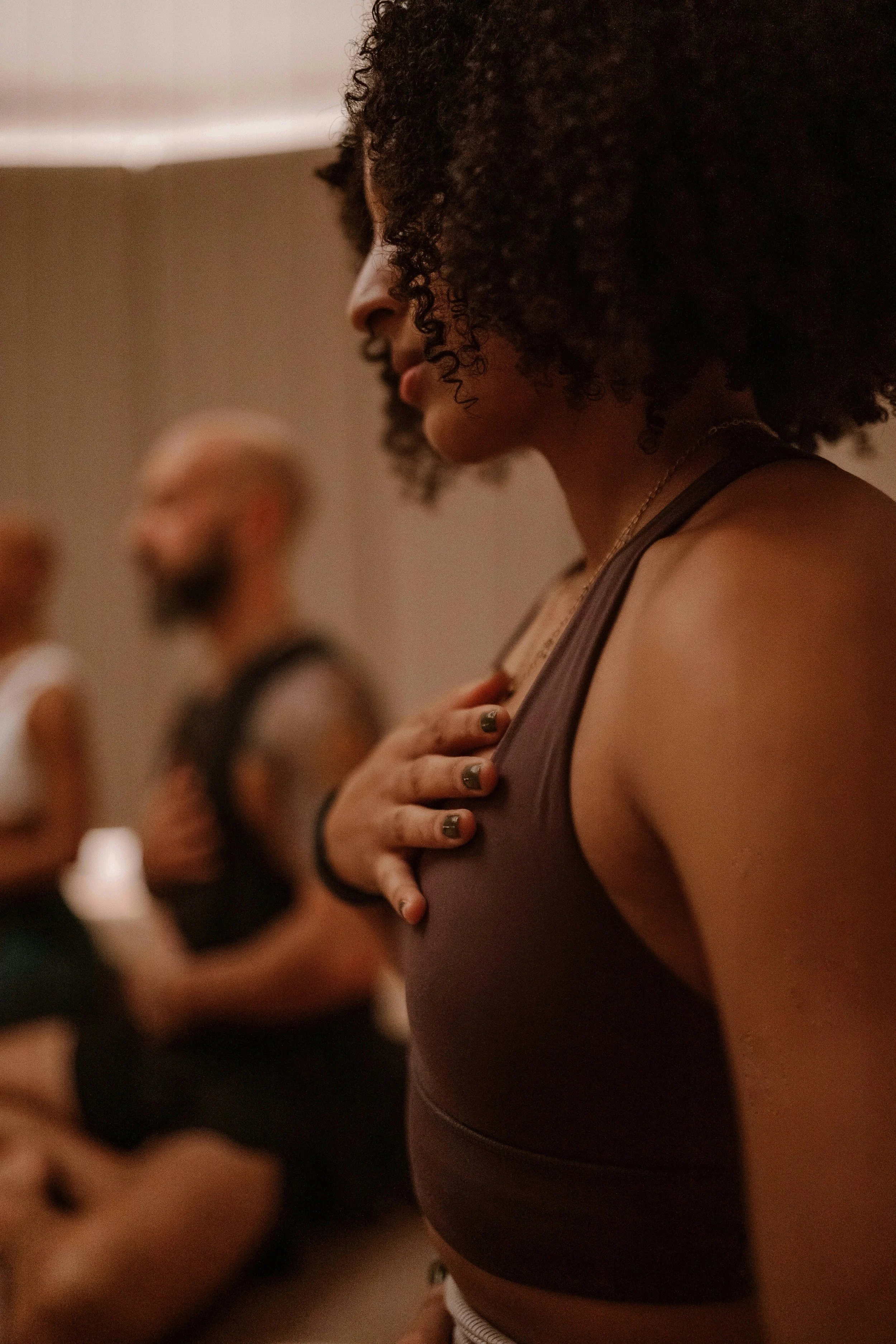 A woman of color, eyes closed, hand resting on her heart, in a group class setting.