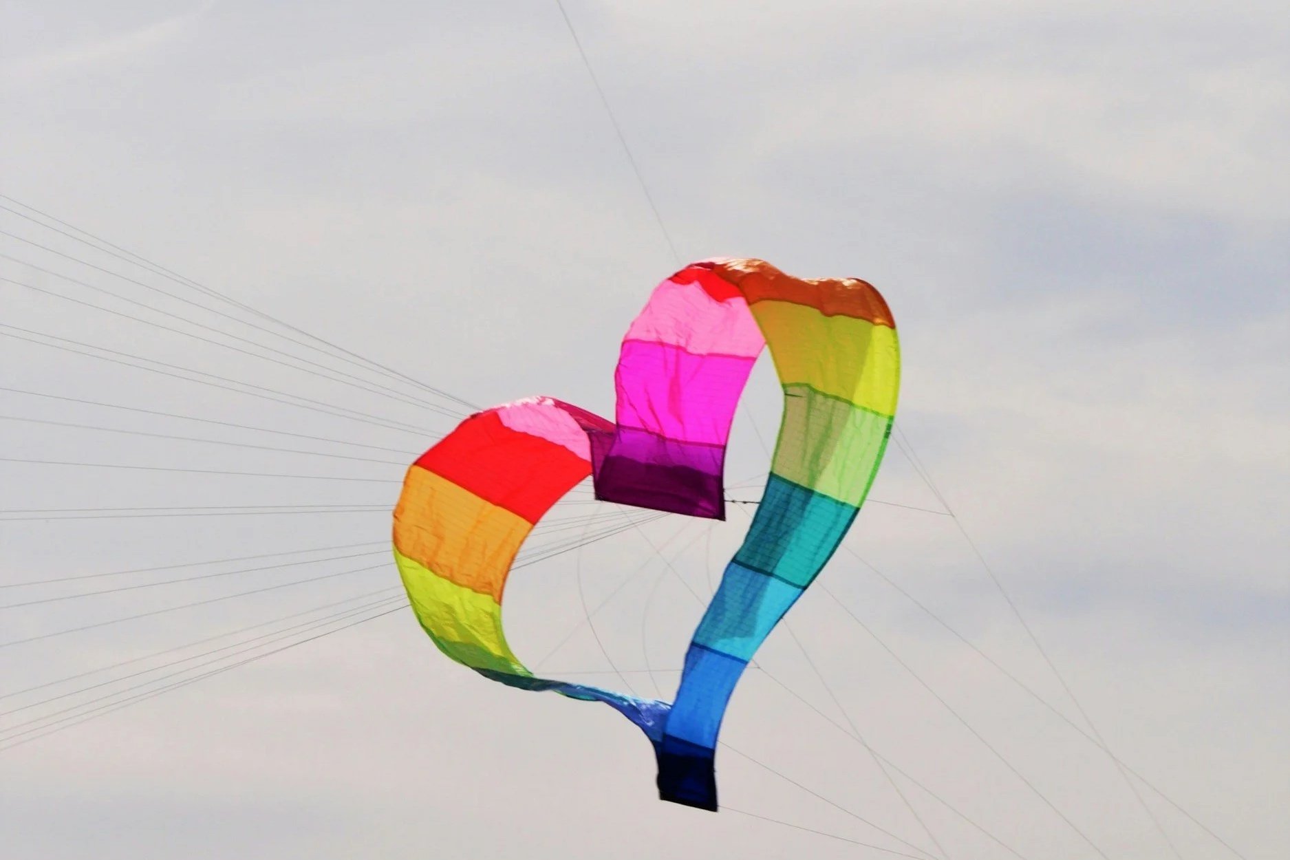 Large rainbow heart kite held aloft in blue sky by multiple strings.