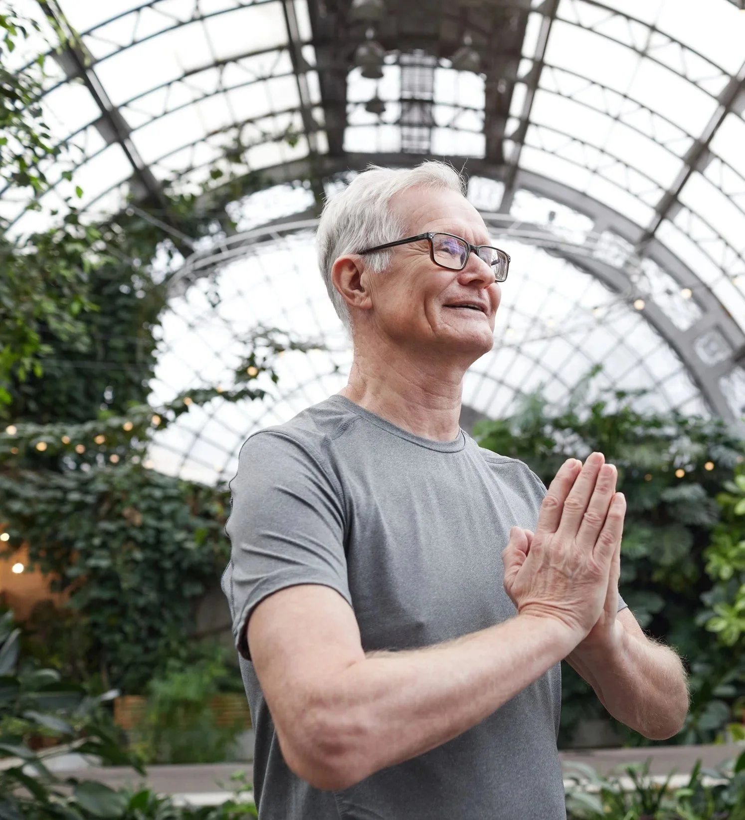 An older man standing in a greenhouse, smiling, his palms meeting at heart centre.