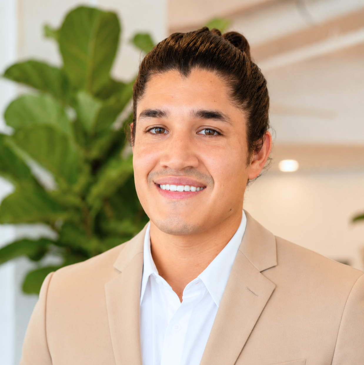 A man with dark hair tied back in a bun, smiling, wearing a beige blazer and white shirt, standing in front of green plants in an indoor setting.