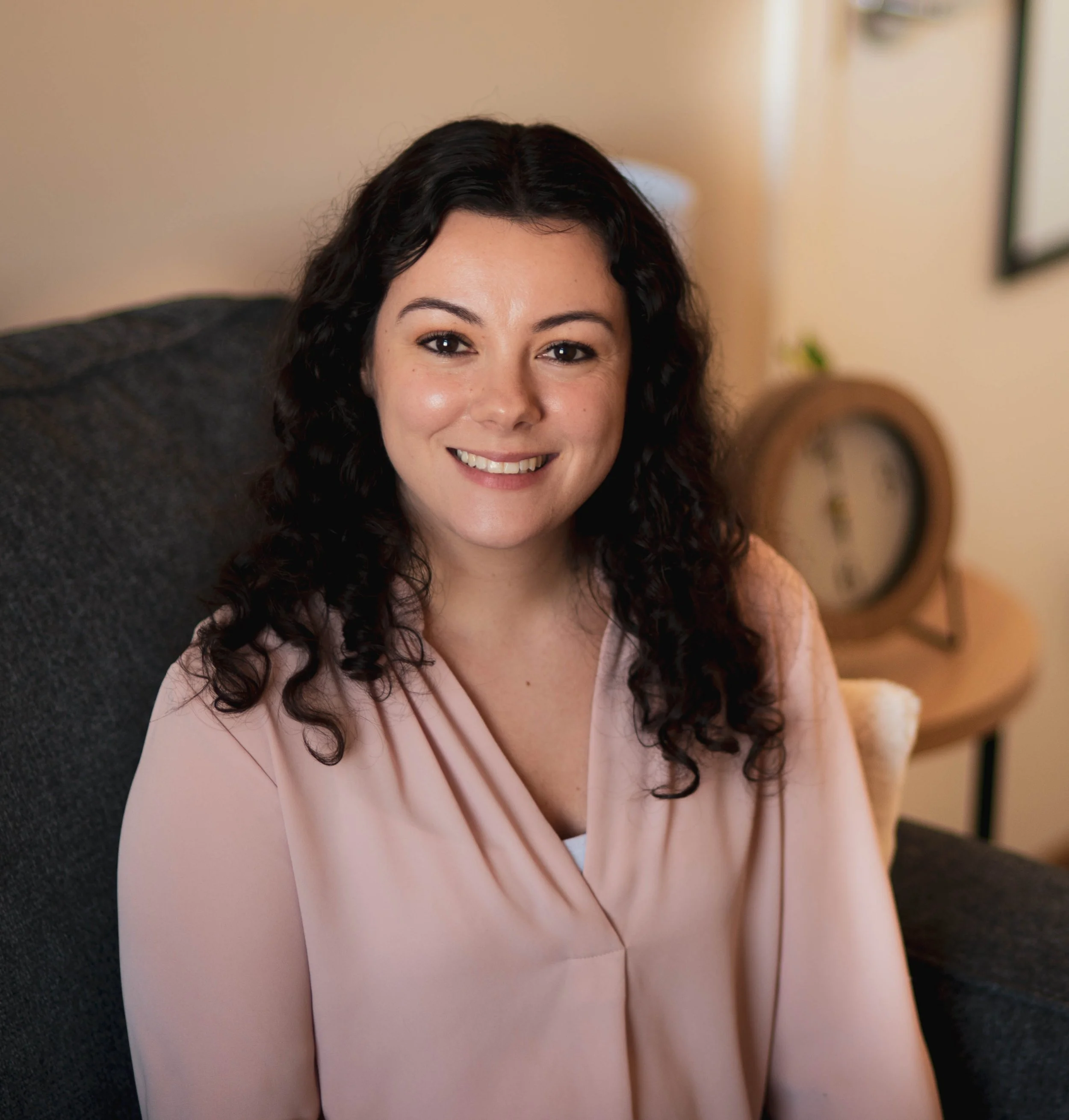 Smiling woman with dark curly hair sitting on a dark gray couch in a cozy room with a clock on a side table in the background.