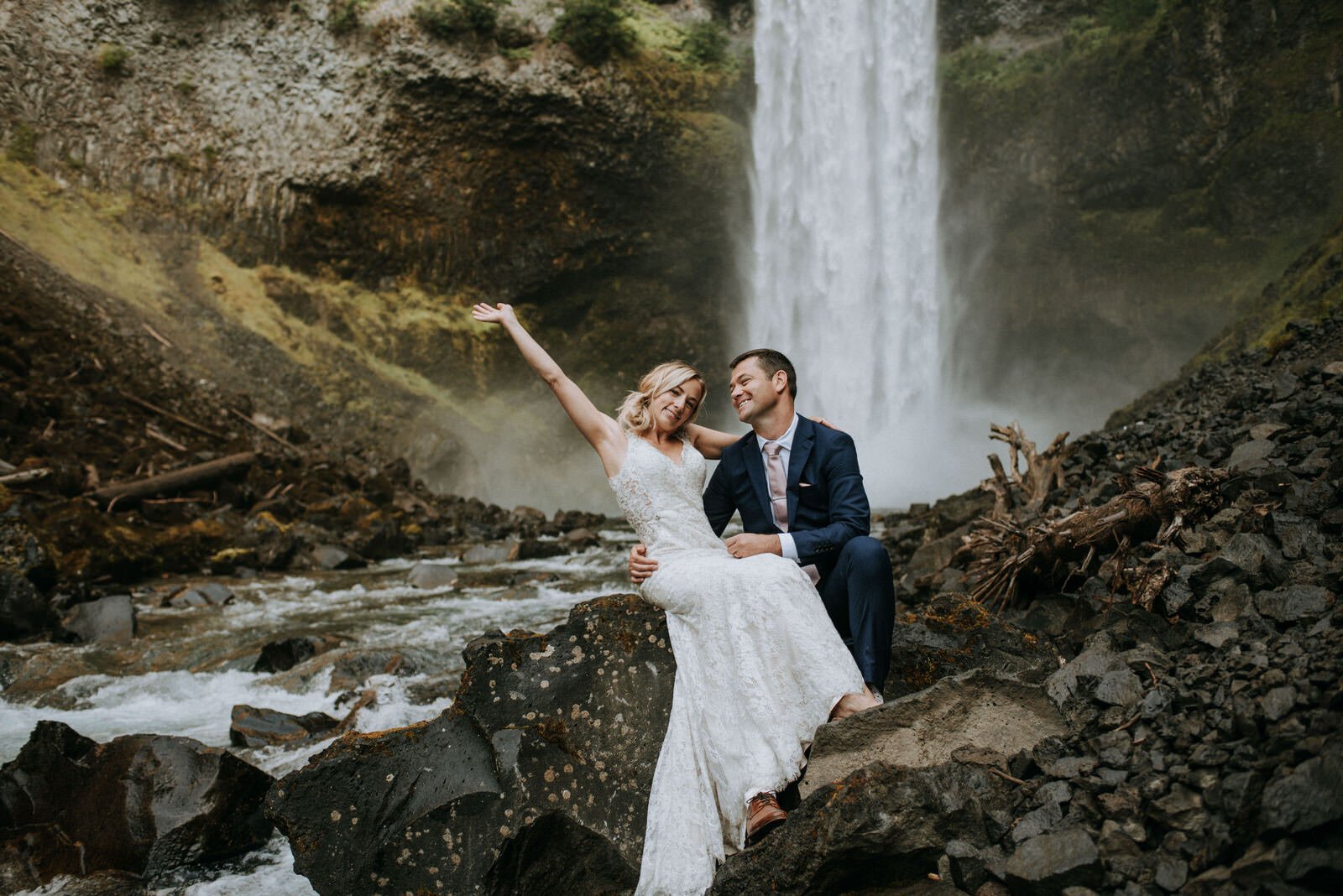 bride sitting on grooms lap in front of waterfall laughing